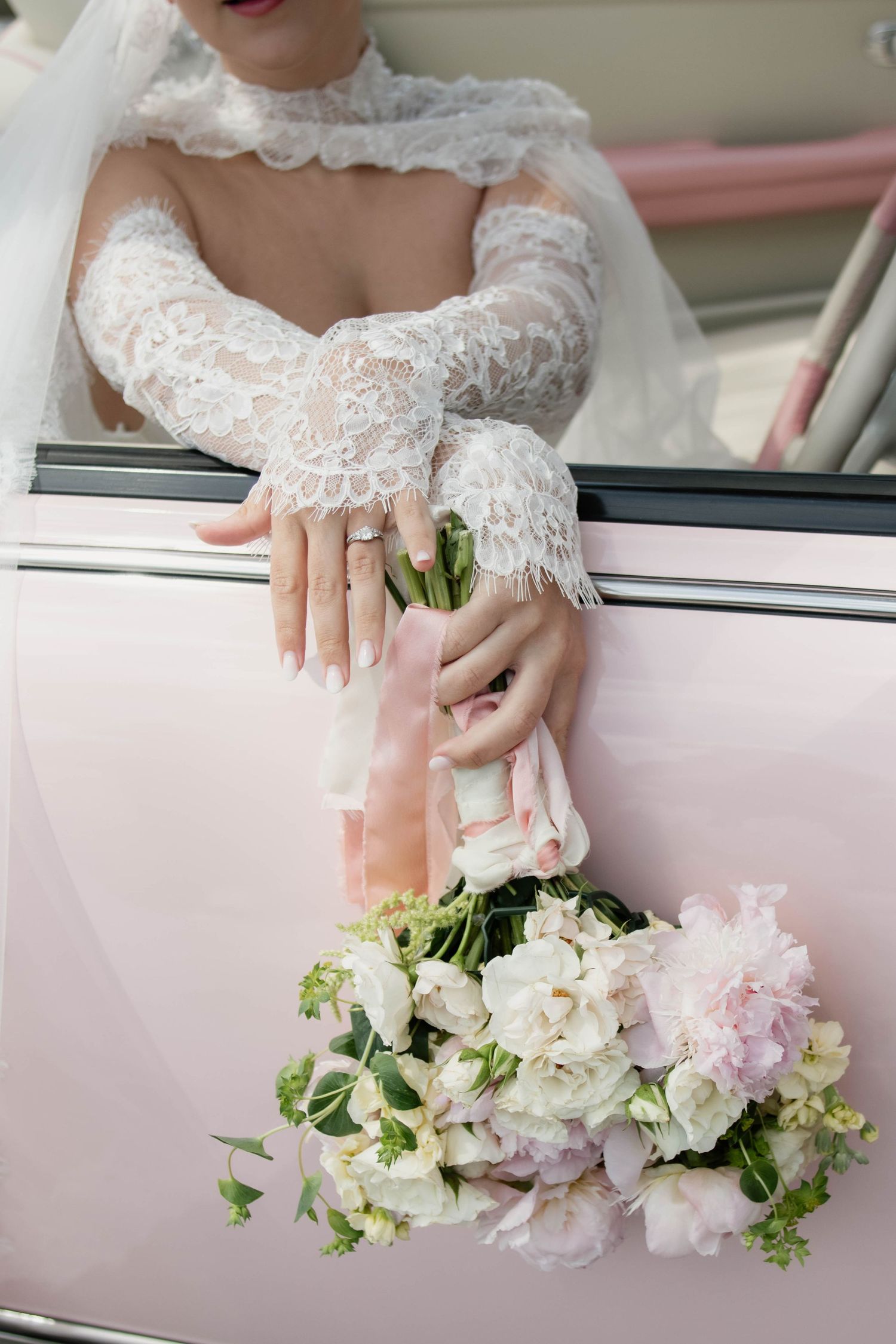 Bride in lace wedding dress holding a bouquet of white and pink flowers out of a pink car window.