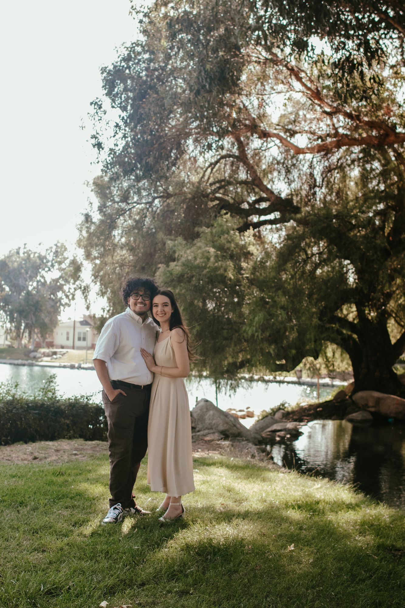 Smiling couple embracing near a pond under a large willow tree in a sunny park.