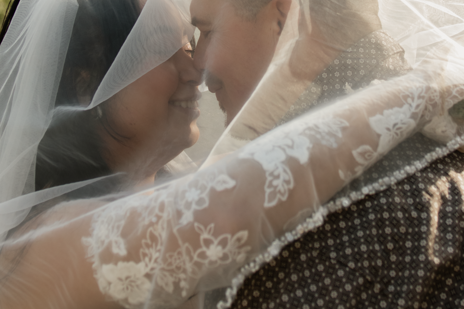 Romantic couple shares intimate moment under flowing lace bridal veil in soft natural light.