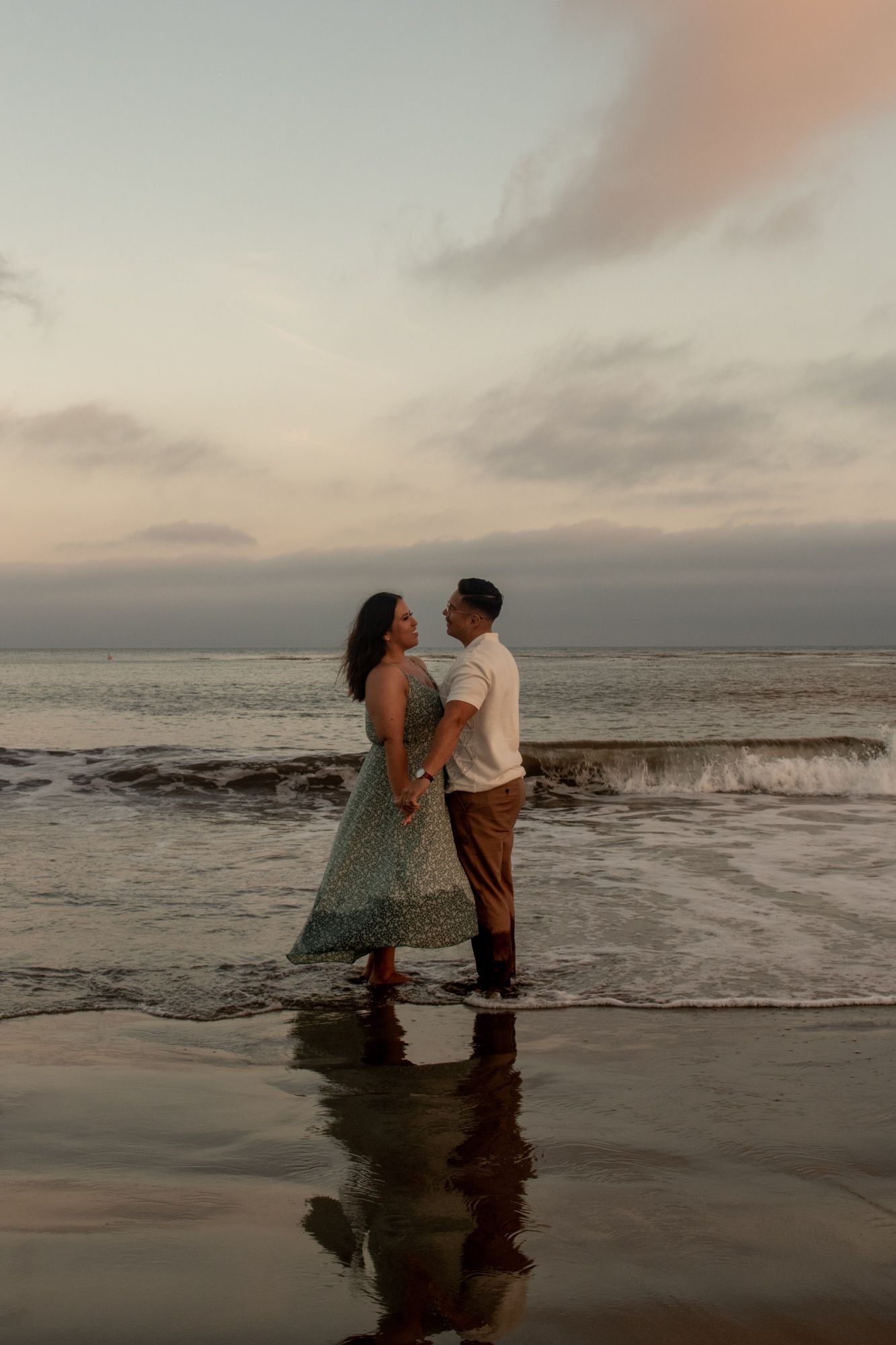 Couple holding hands facing each other on beach at sunset with ocean waves behind them.