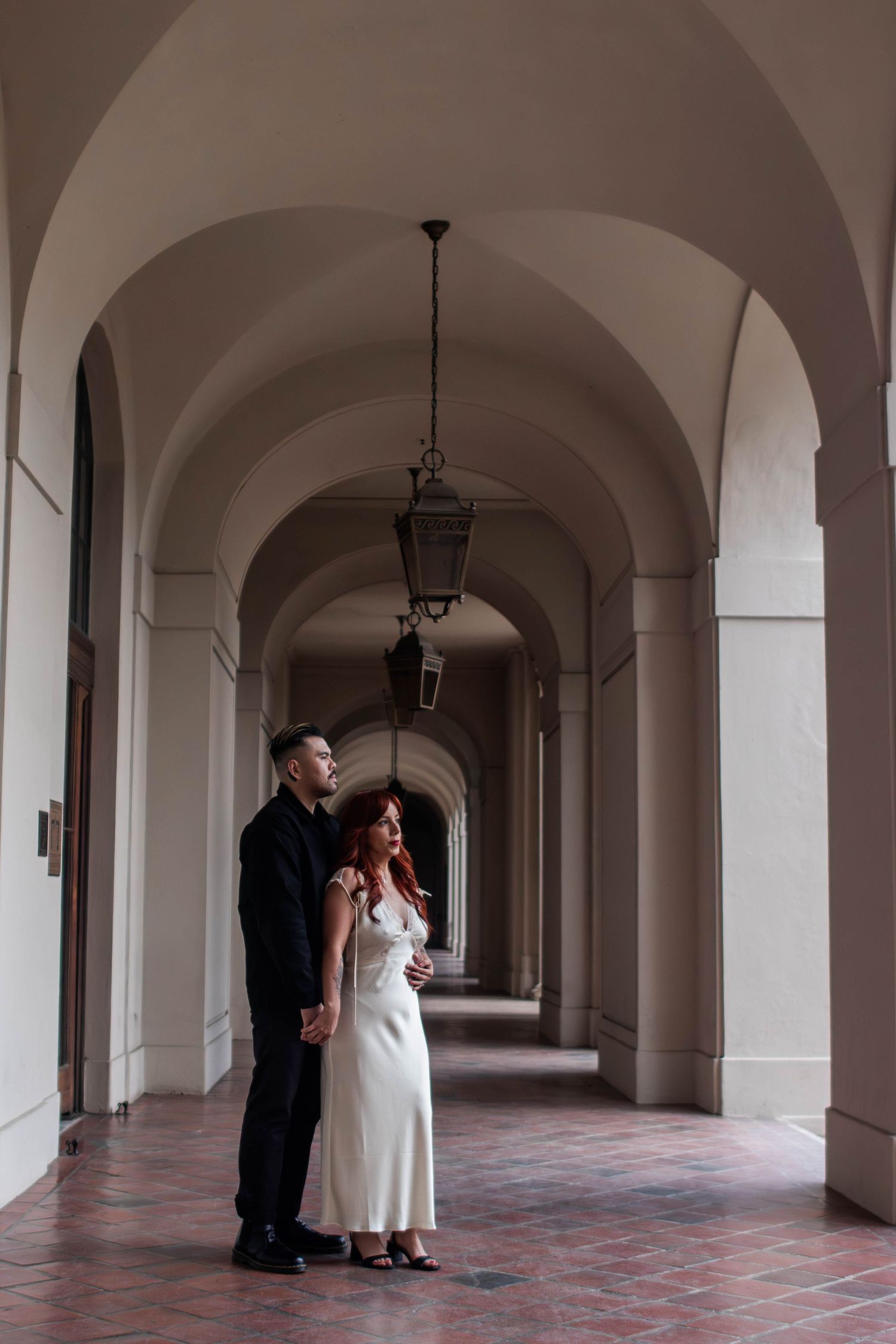 Couple standing together in a long arched corridor with hanging lanterns and terracotta tile floor.