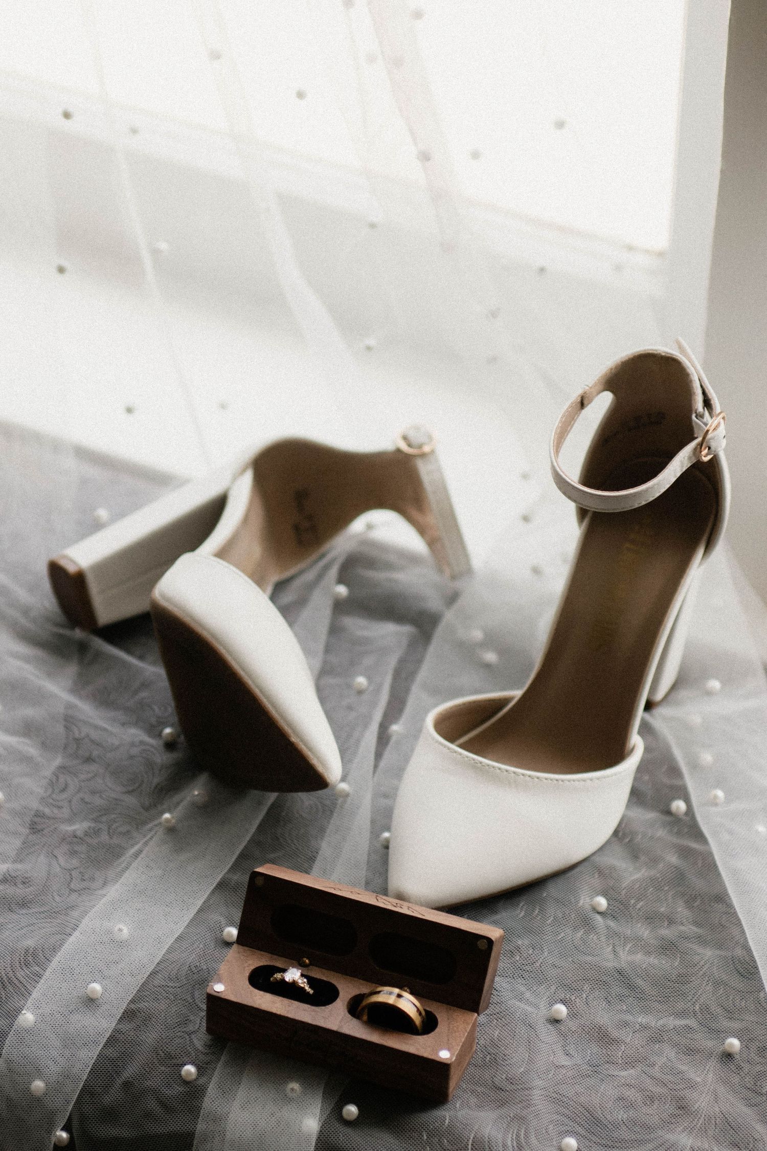 White bridal heels with ankle straps beside a wooden ring box holding two gold wedding bands.