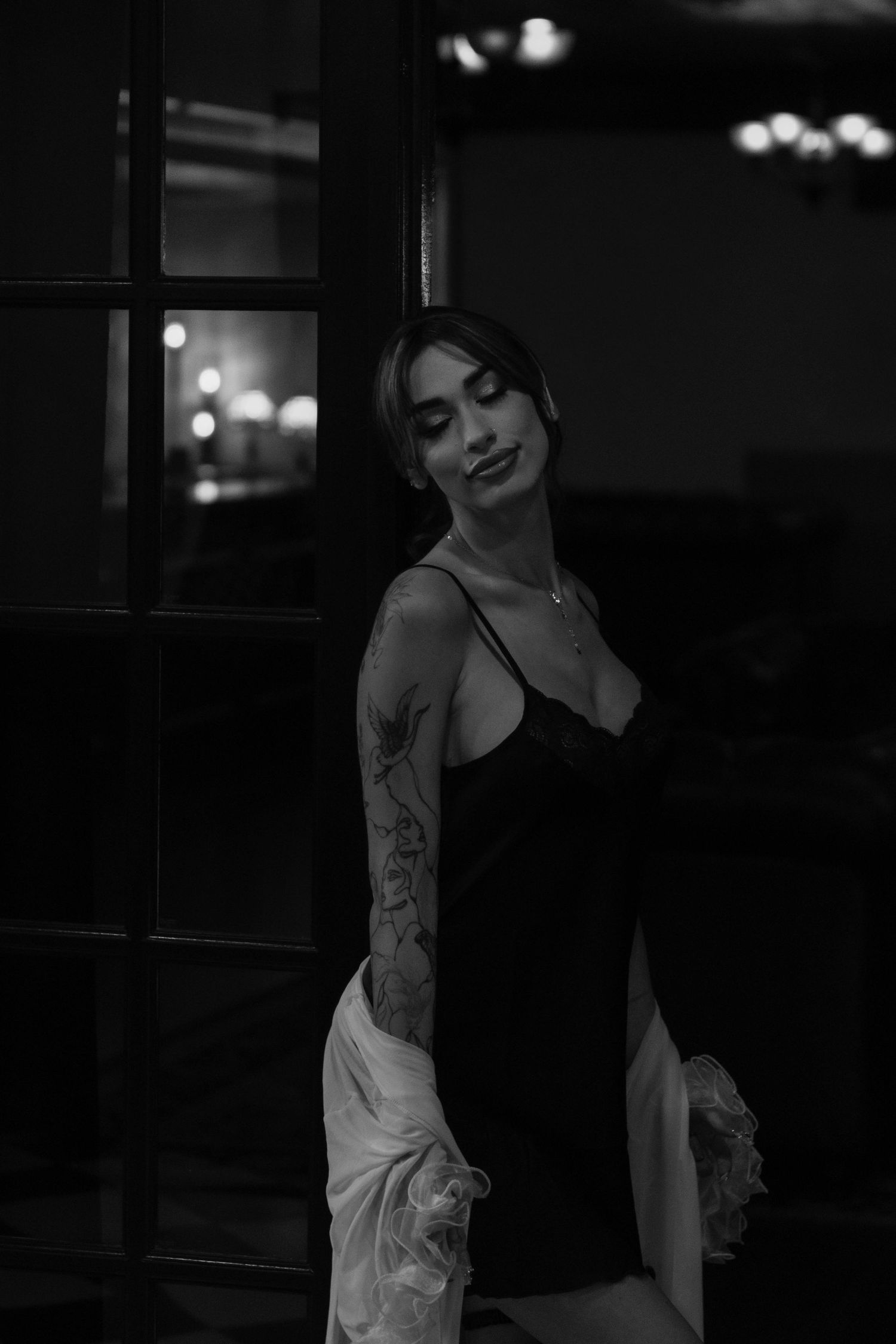 Black and white portrait of a tattooed woman in a black dress posing elegantly near a dark doorway.