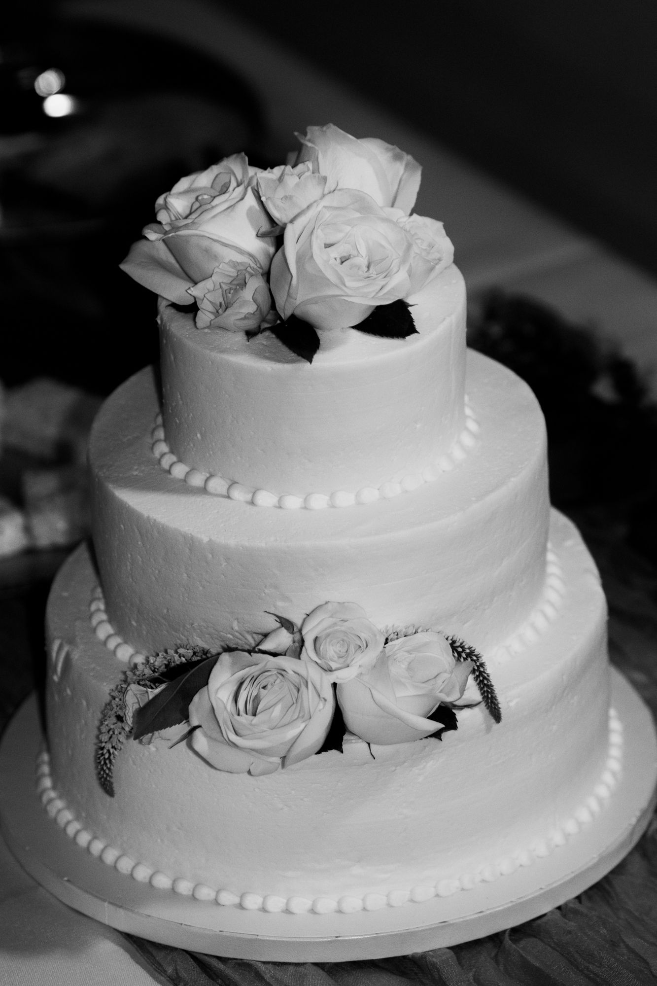 Elegant three-tier white wedding cake decorated with roses and pearl borders in black and white photography.