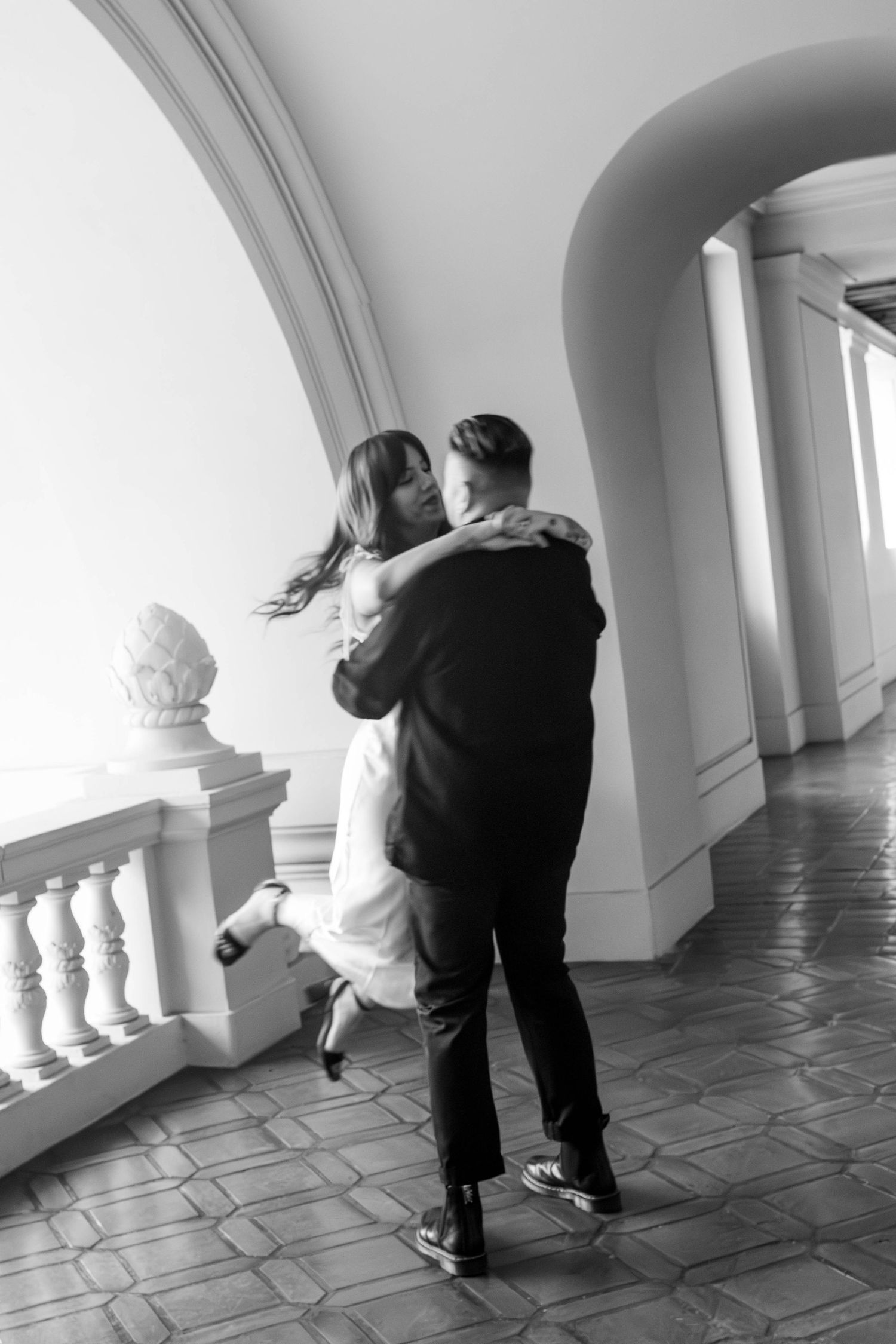 Black and white photo of a groom lifting his bride in a grand arched hallway with balustrades.