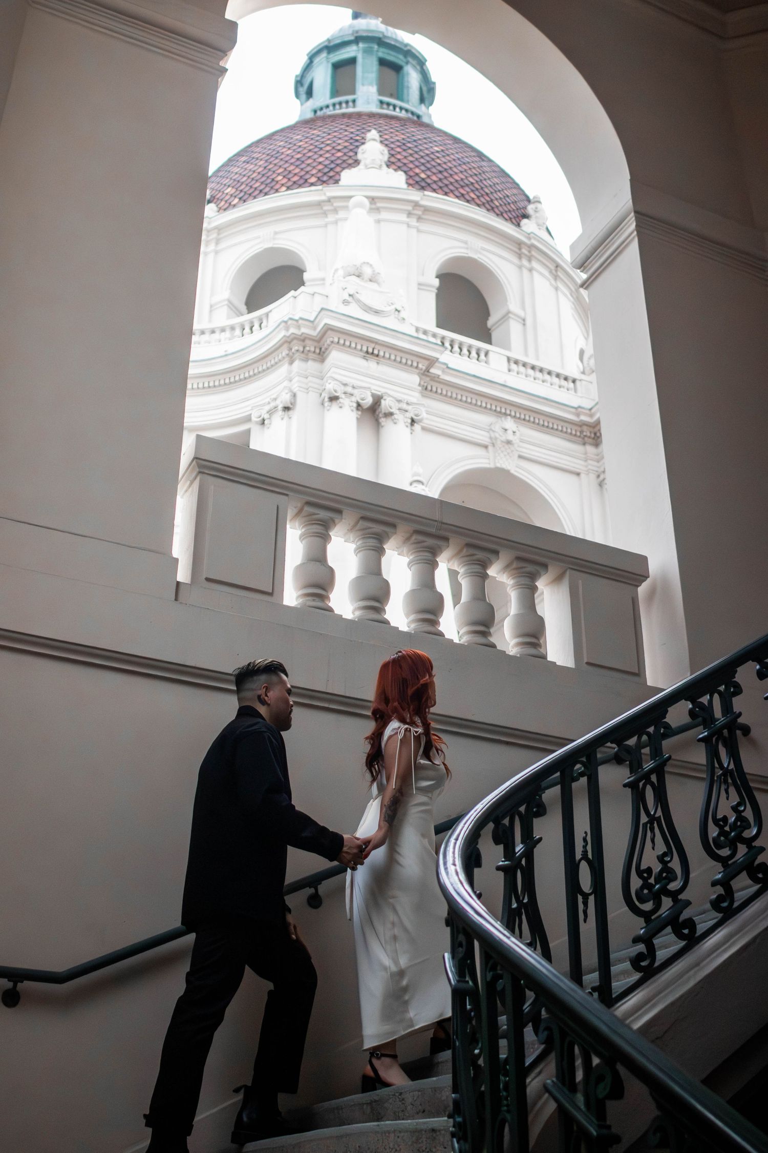 Couple holding hands ascending grand staircase beneath ornate domed building archway.