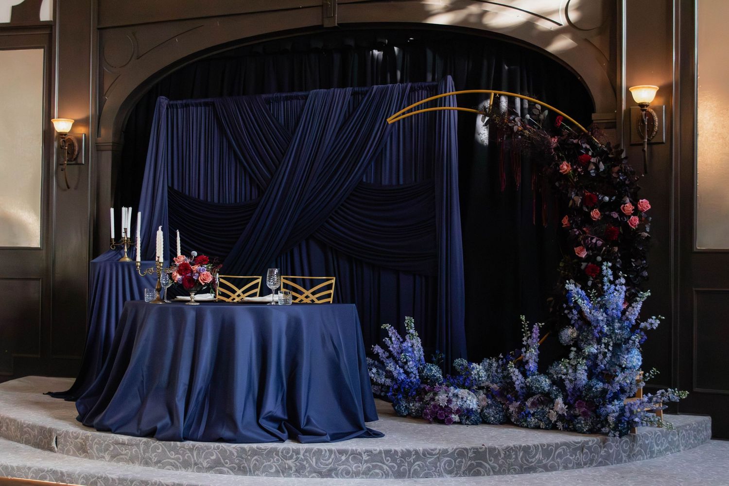Elegant navy blue wedding sweetheart table with floral arch, candles, and blue flower arrangements on a raised stage.