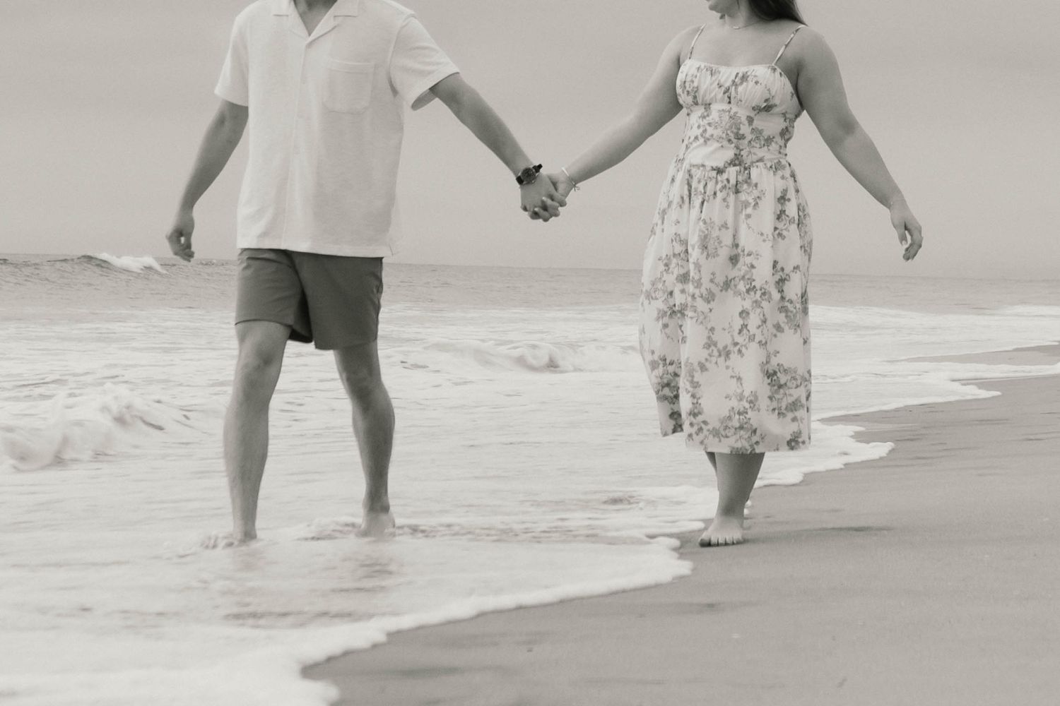 Black and white photo of couple holding hands walking along shoreline with ocean waves.