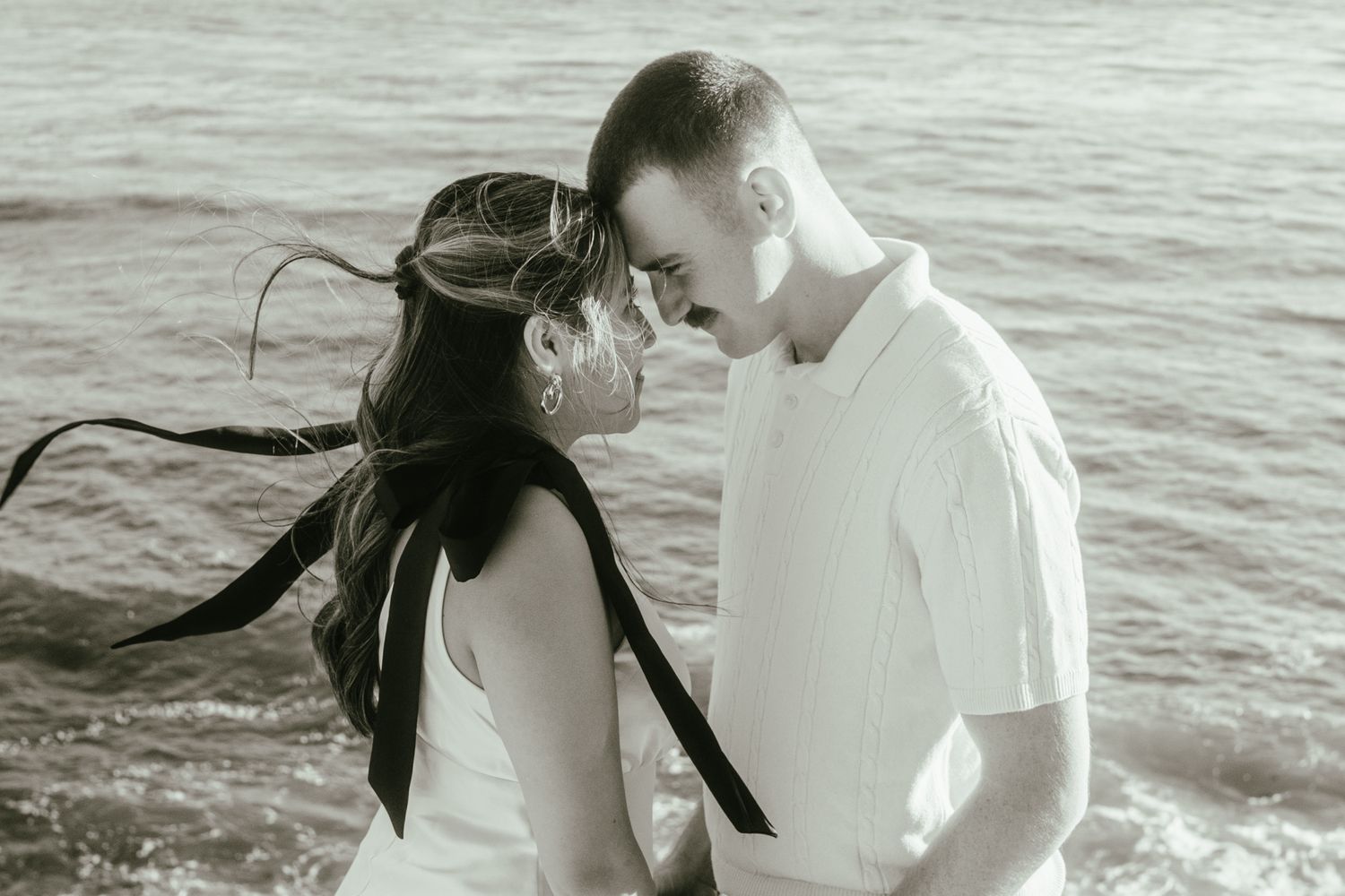 Romantic black and white beach photo of couple nearly kissing with wind-blown hair by the ocean.