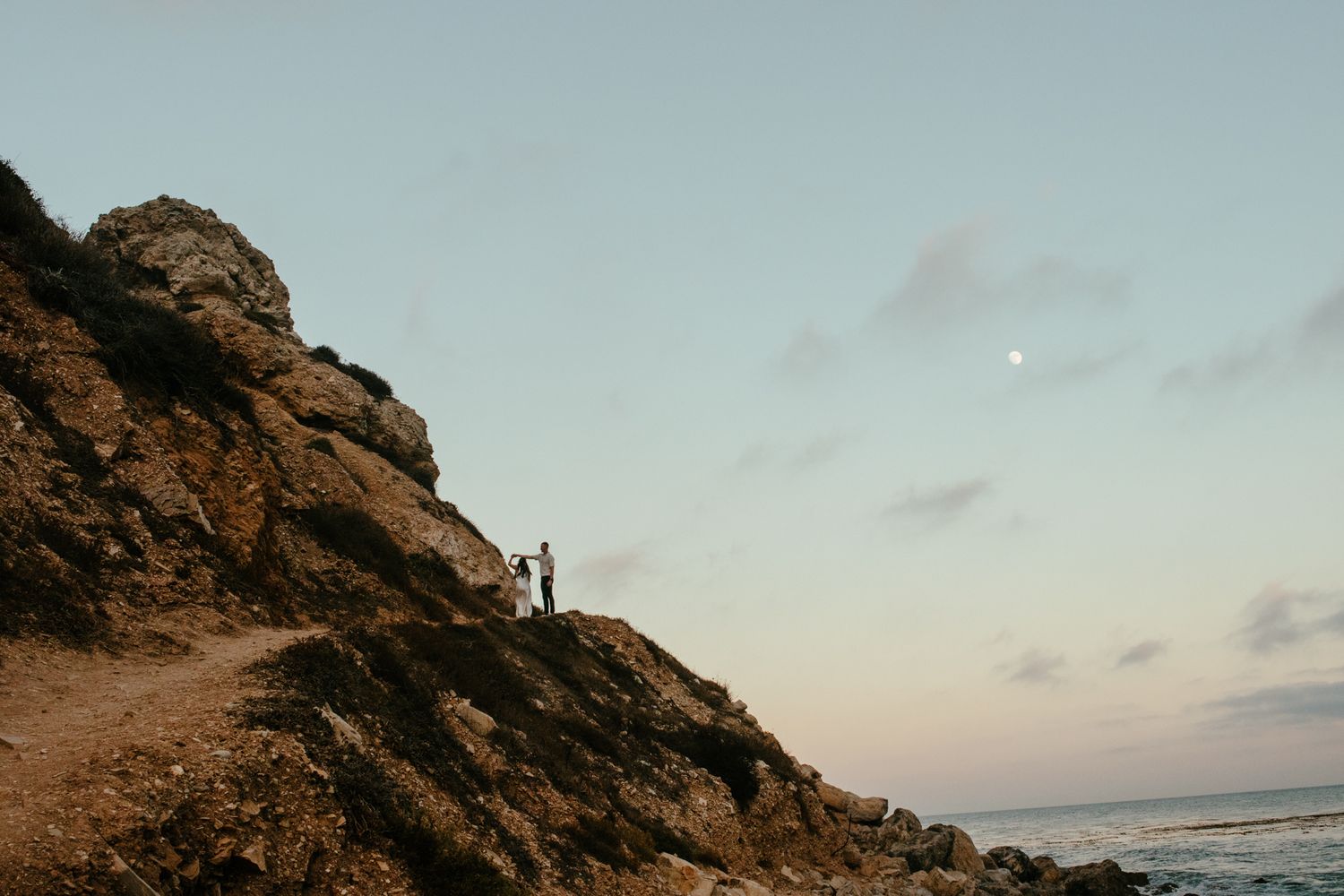 Two figures stand on dramatic rocky coastal cliffside trail at twilight with crescent moon rising.
