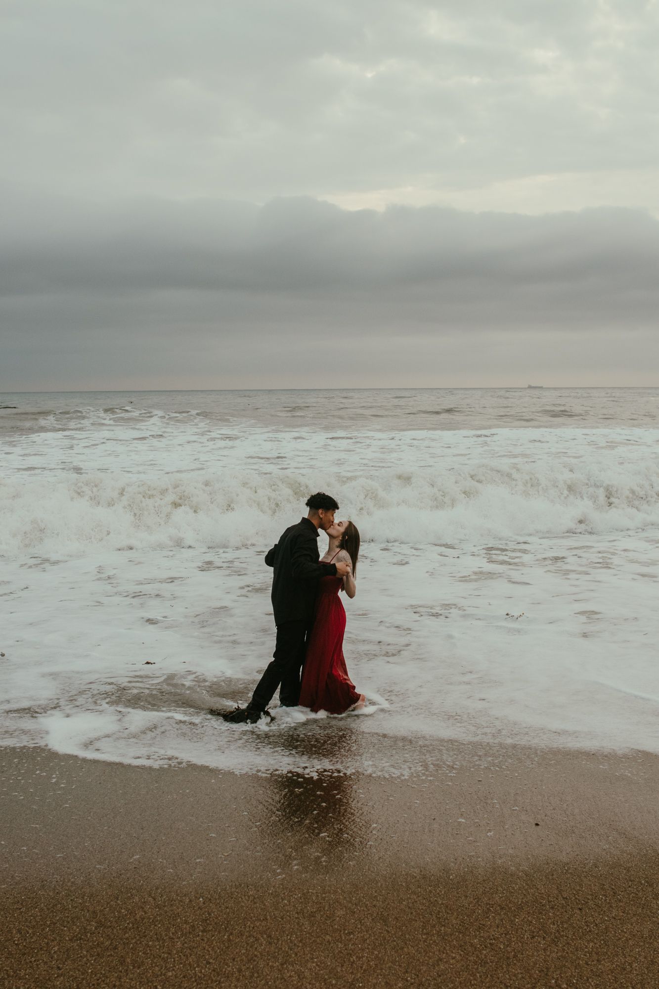 Couple kissing in crashing ocean waves on stormy beach, woman wearing red gown and man in black suit.