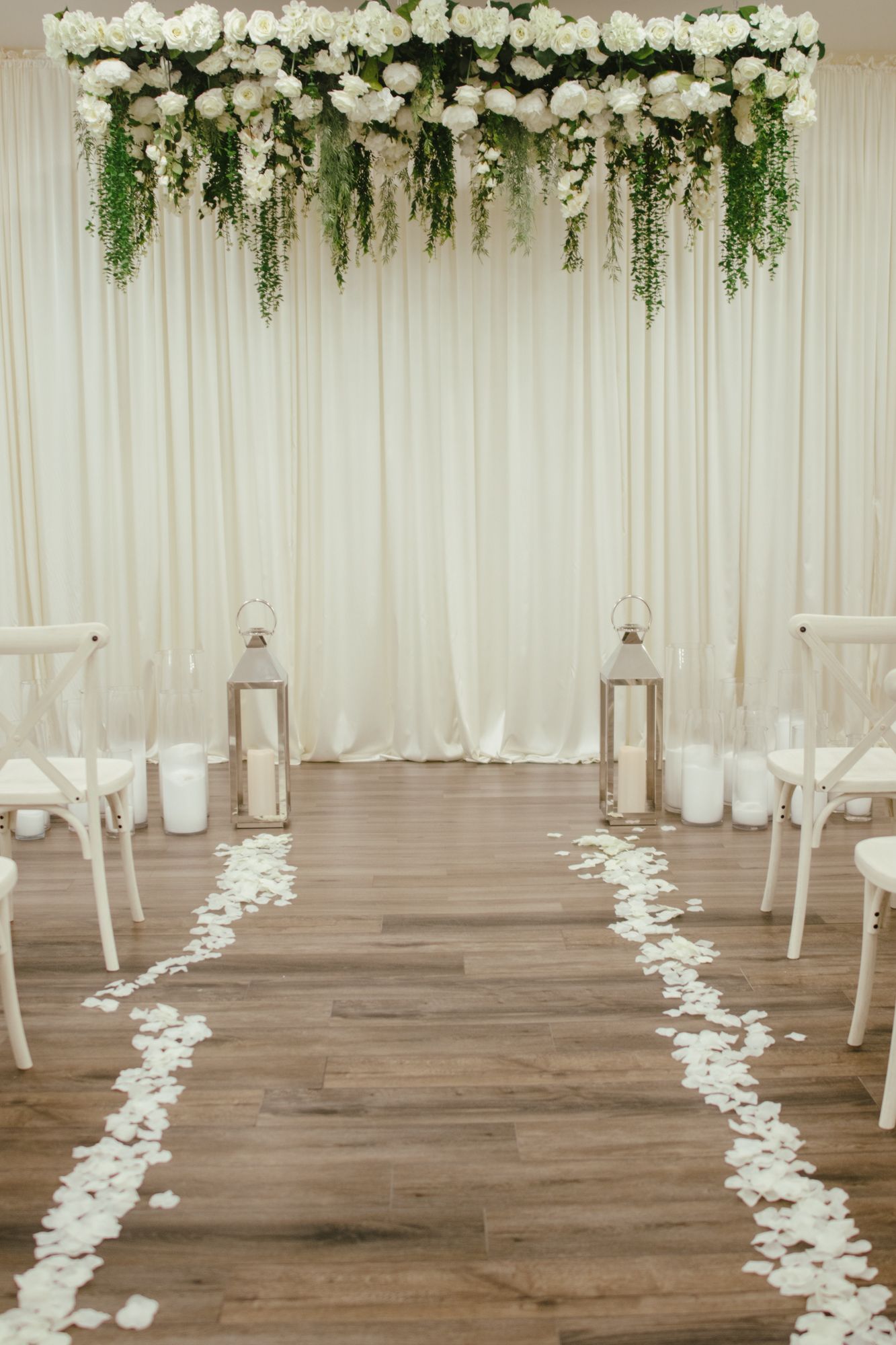 White floral arch with rose petal aisle and lanterns at an indoor wedding ceremony.