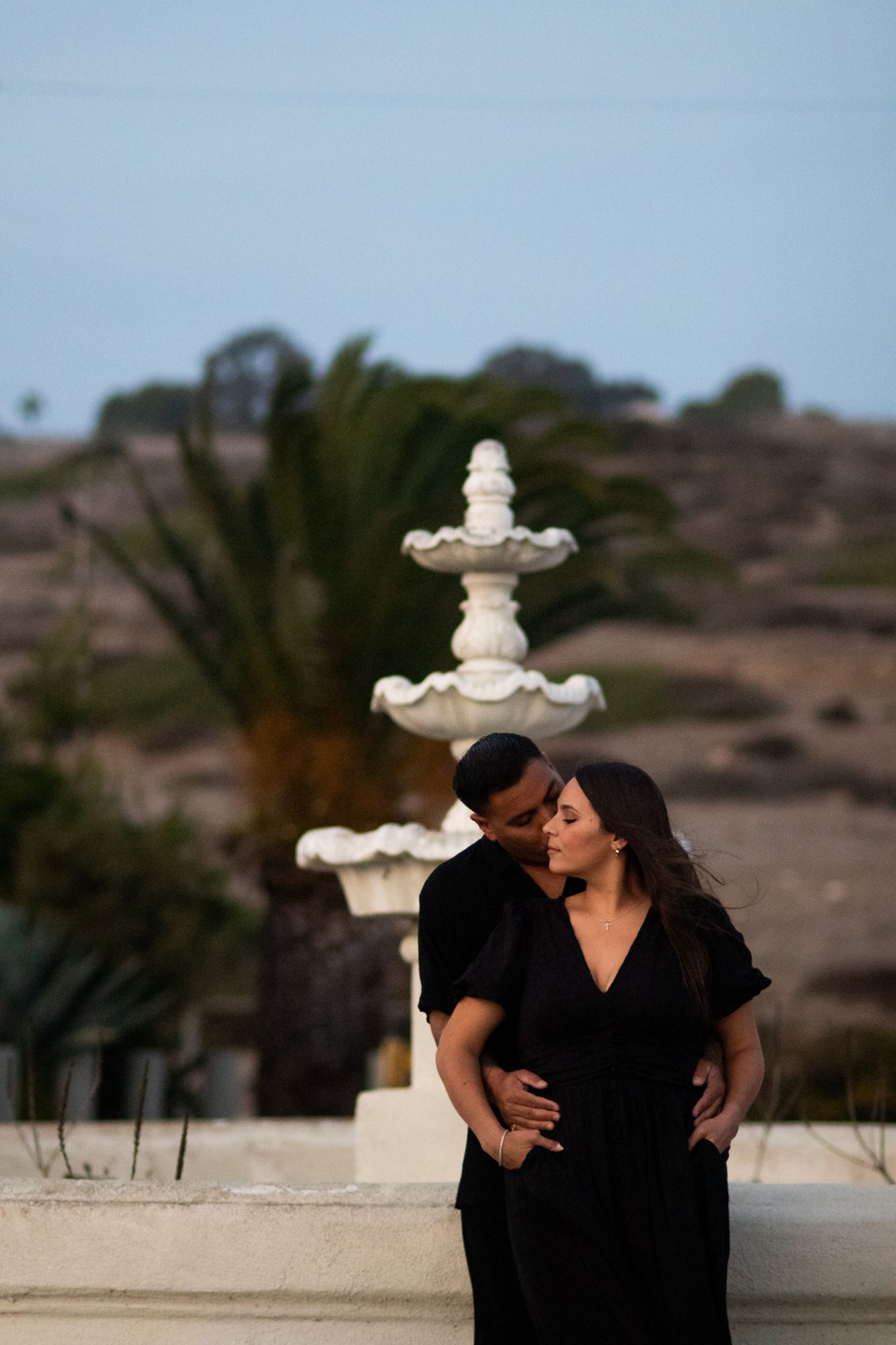 Couple in matching black outfits embracing near a white stone fountain with palm trees at dusk.