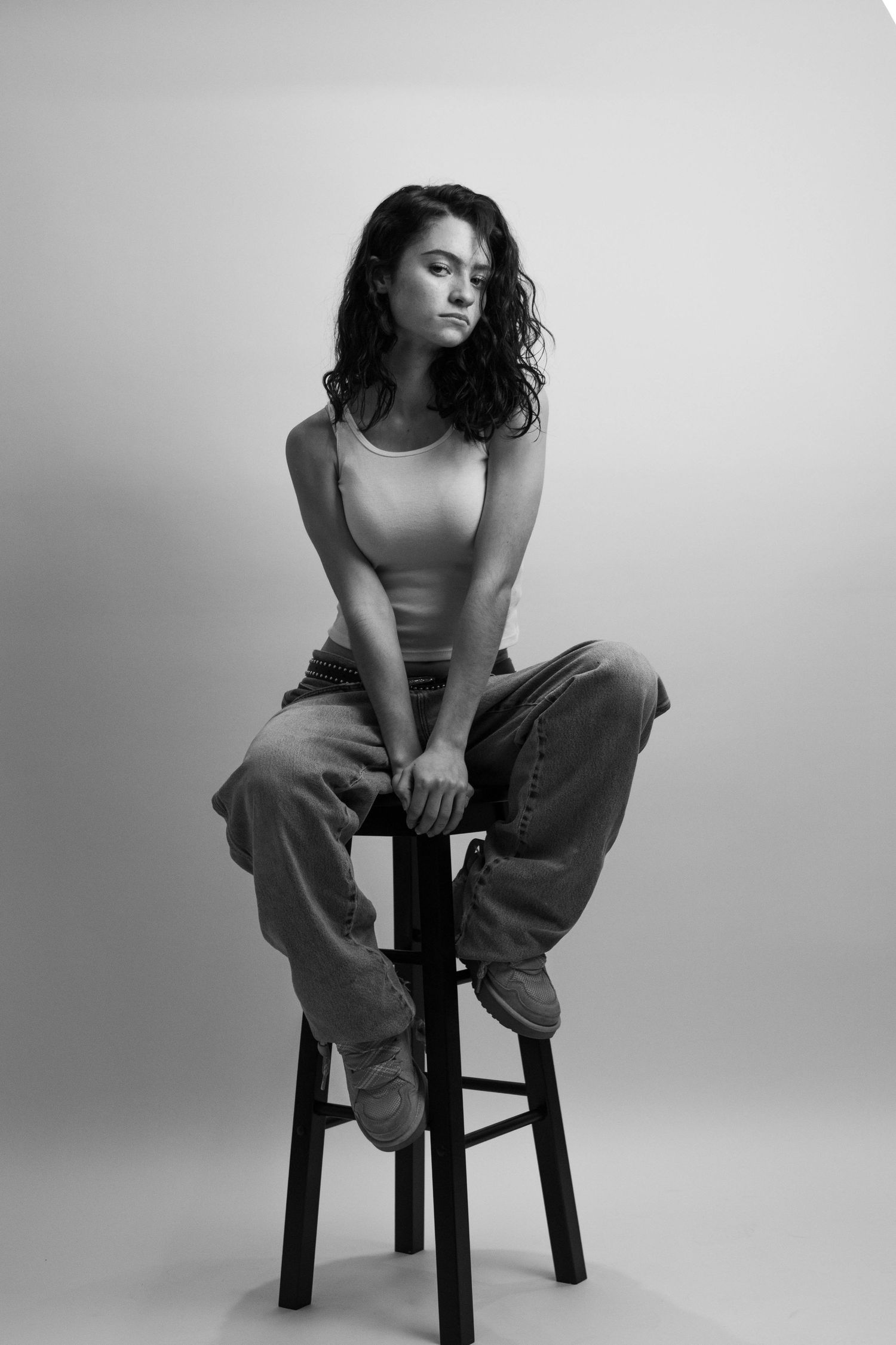 Black and white portrait of woman sitting on stool in tank top and jeans.