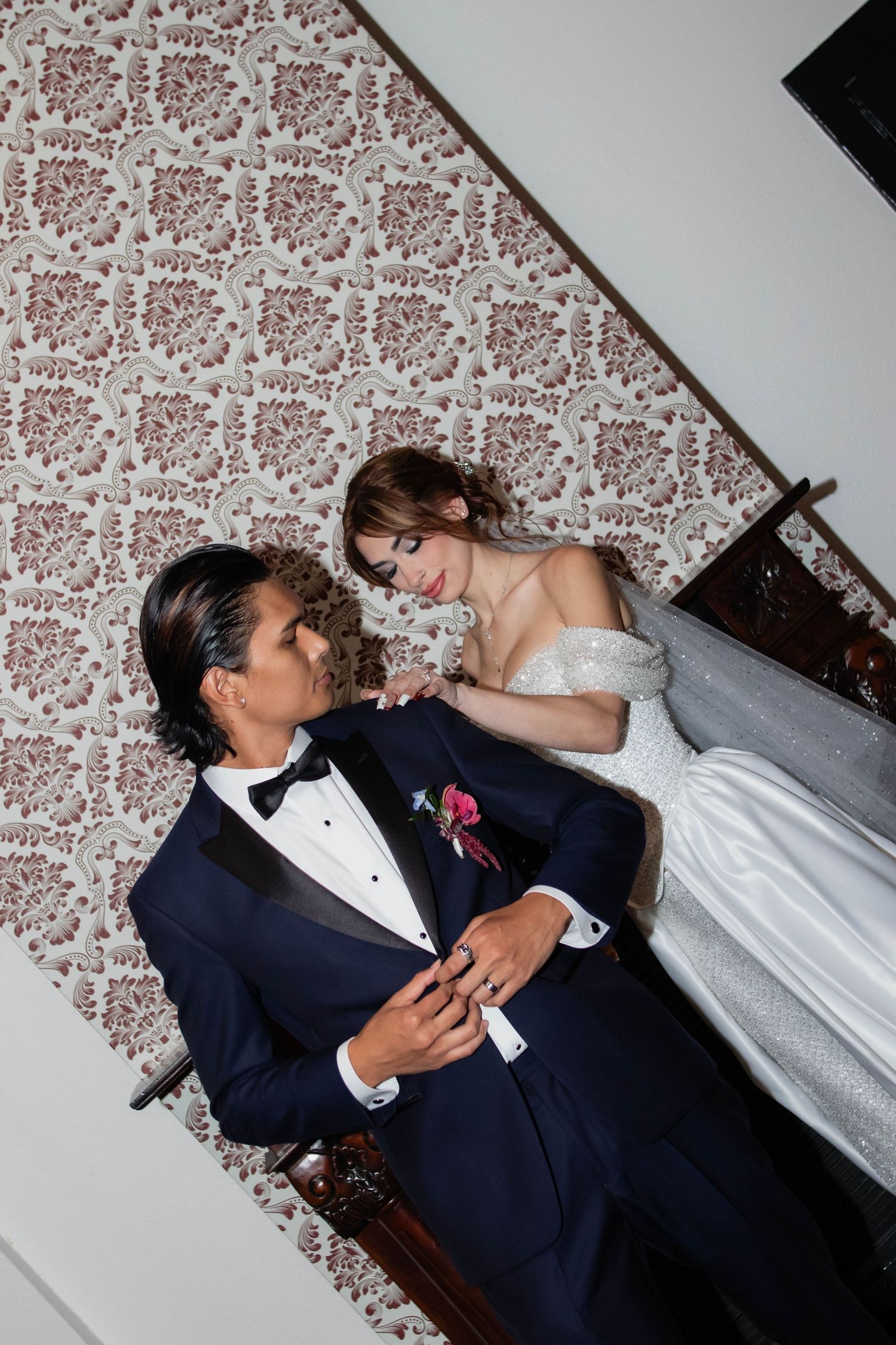 Bride in white gown and groom in navy tuxedo share a romantic moment on a staircase with floral wallpaper.