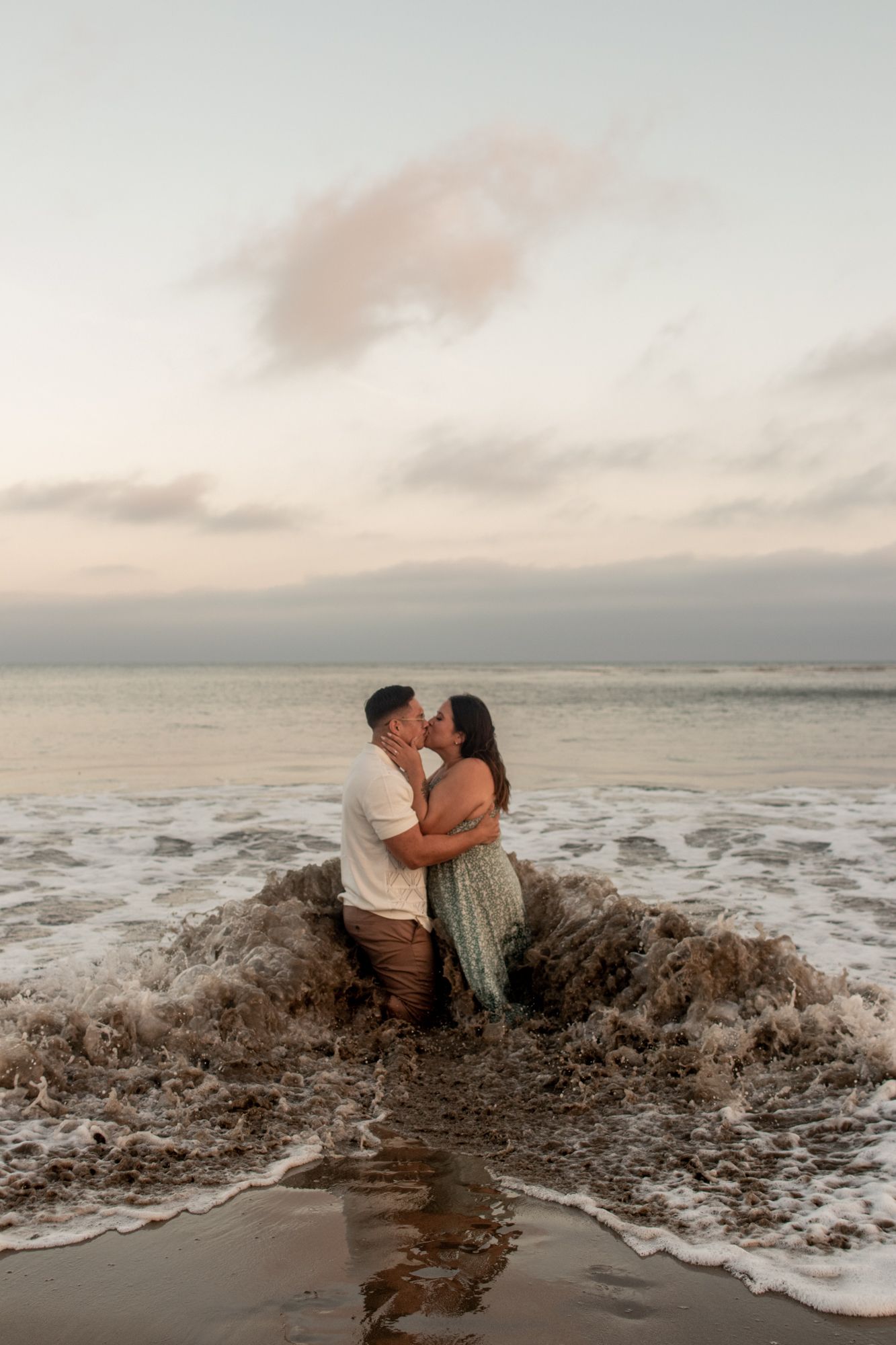 A couple kisses romantically while standing in ocean waves on a beach at sunset.