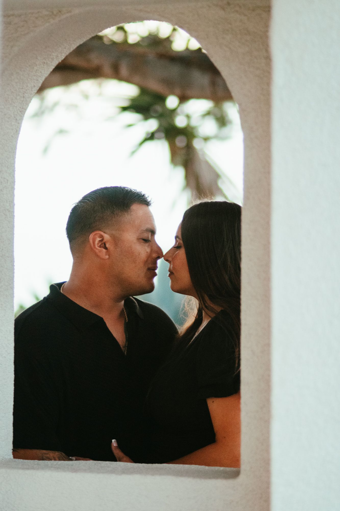 Couple in black outfits sharing an intimate moment framed by a white arched window opening.