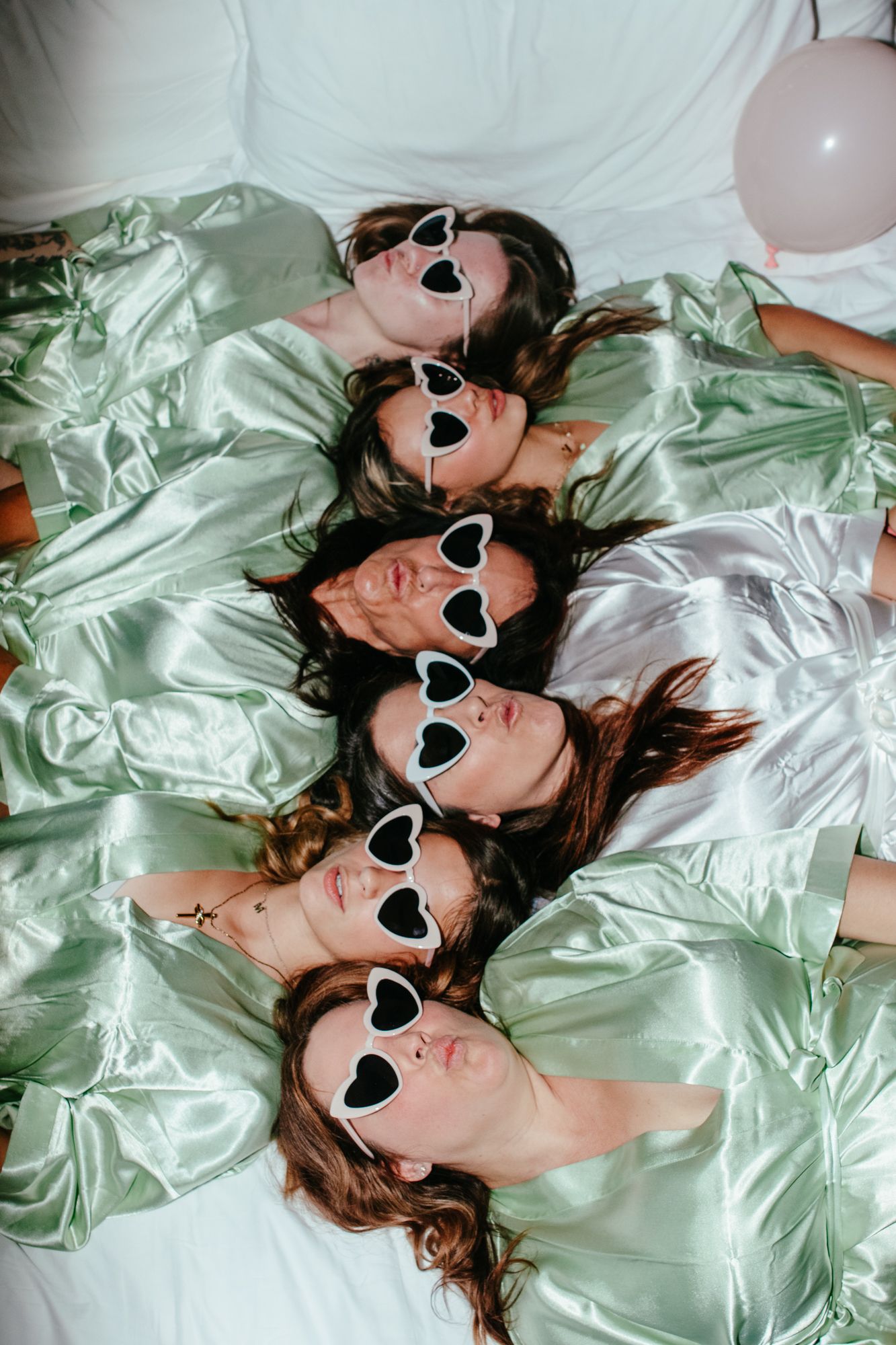 Five women in satin robes lying together wearing heart-shaped sunglasses at a bachelorette celebration.