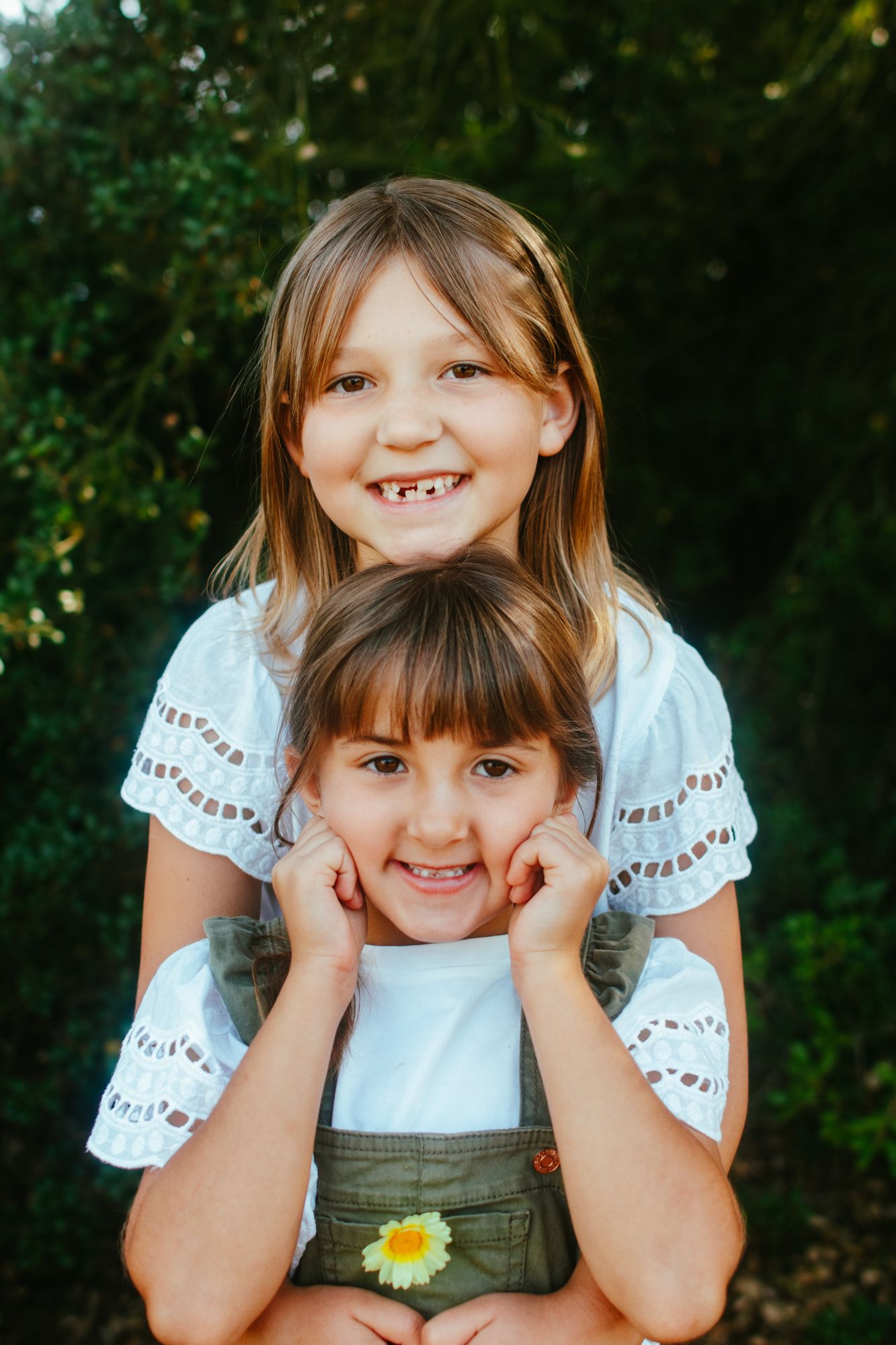 Two smiling young sisters posing together outdoors with green foliage background.