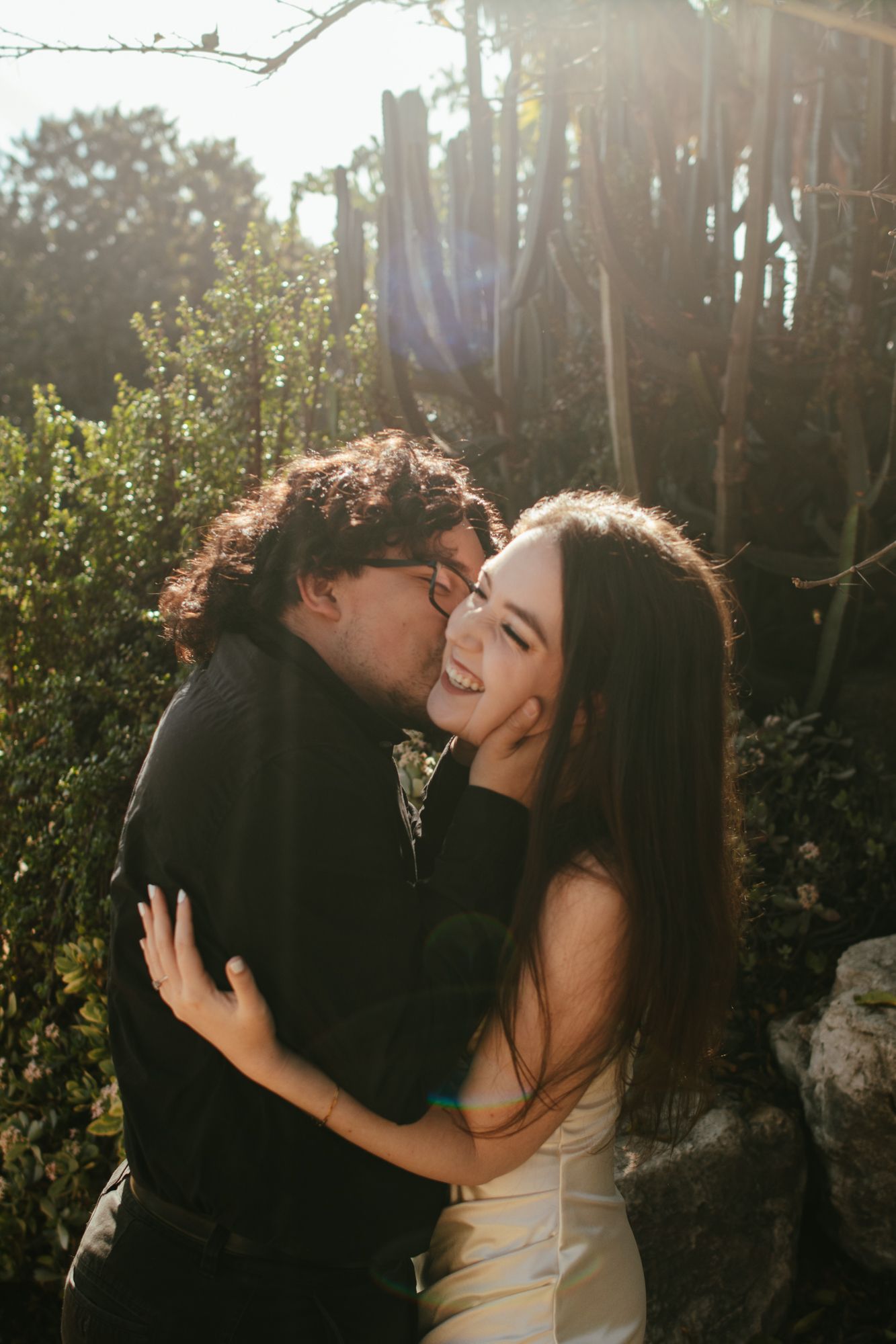 Joyful couple embracing in sunlit garden near cacti, man kissing laughing woman's cheek outdoors.