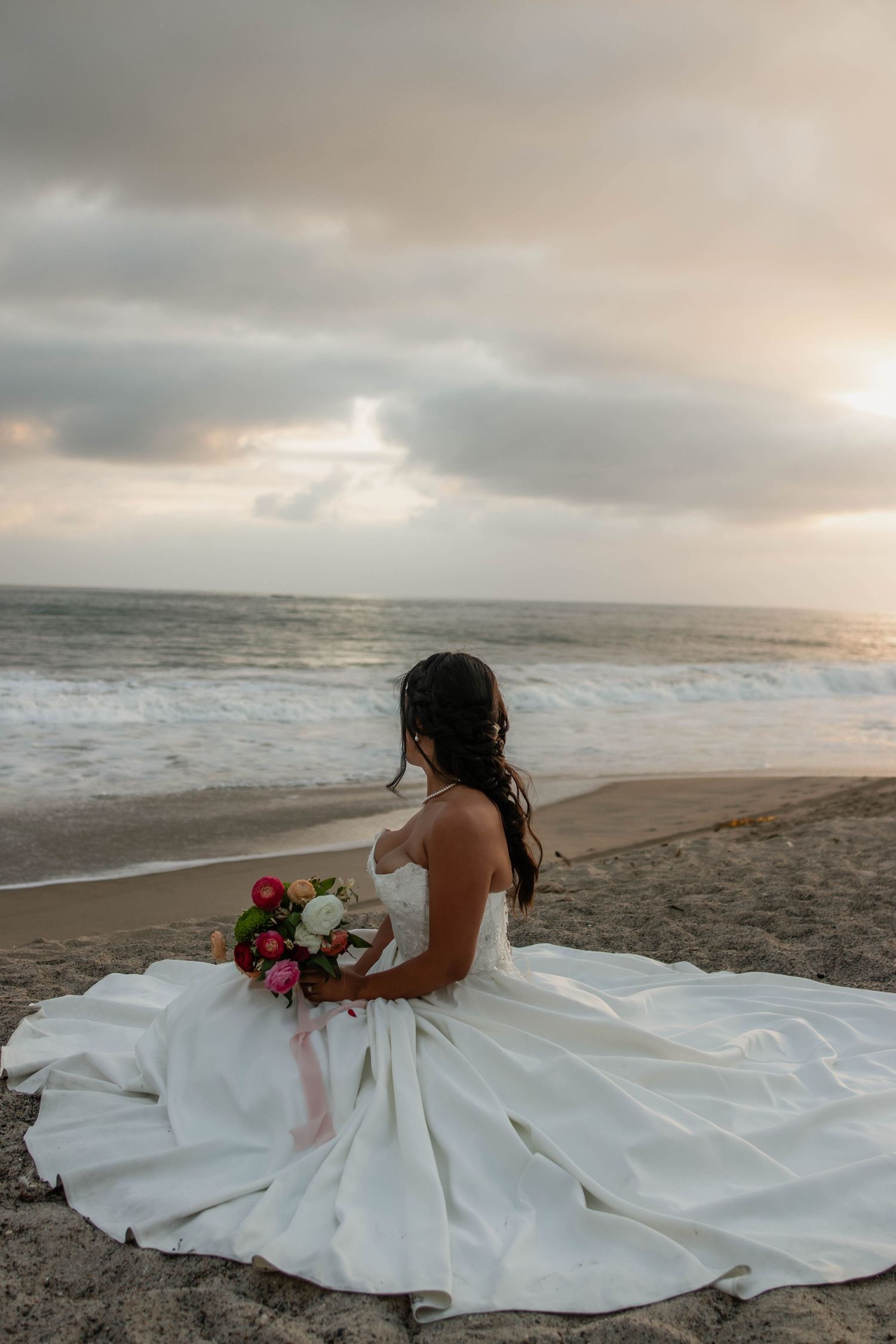 Bride in white gown sitting on beach with colorful bouquet, gazing at ocean waves during cloudy sunset.