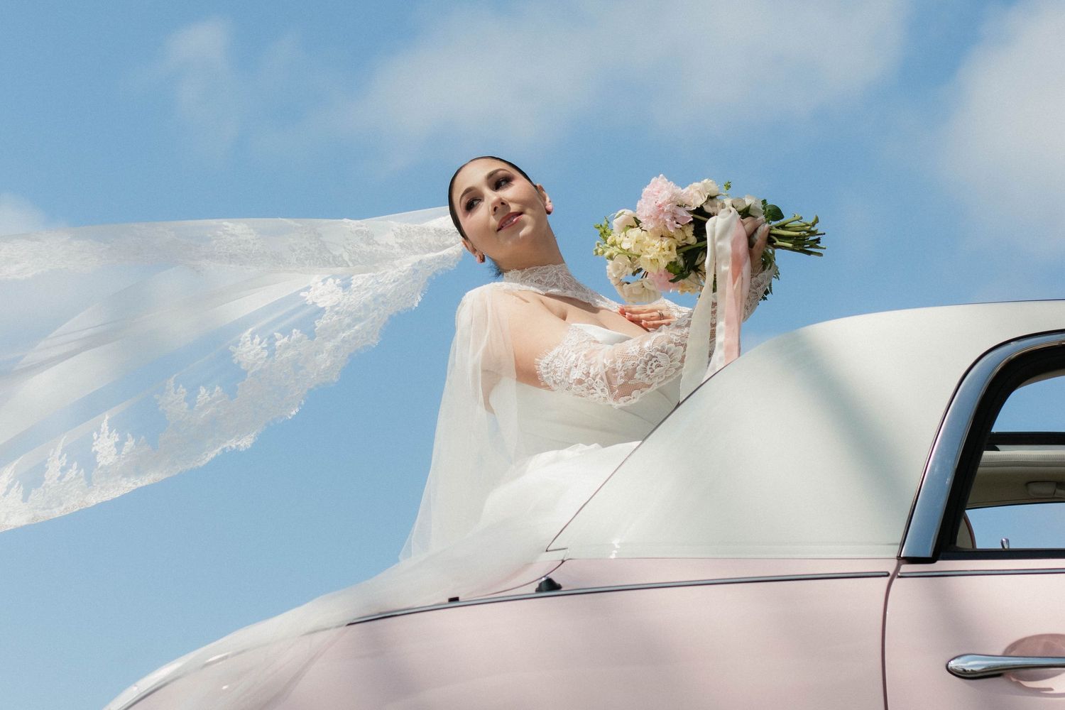 Bride in white gown and veil holds bouquet while leaning out of a vintage car under a blue sky.