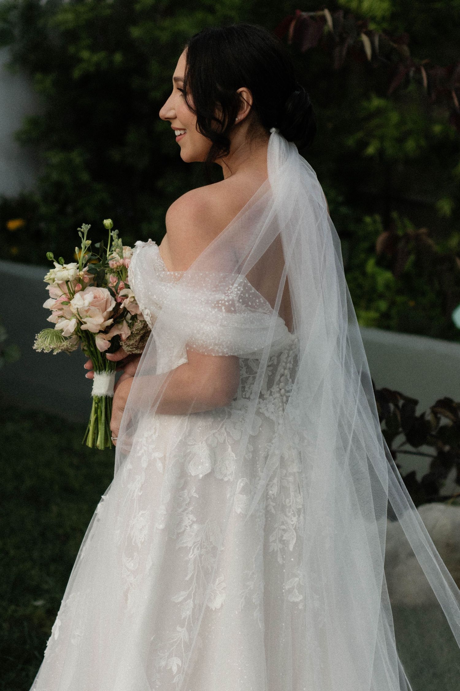 Bride in off-shoulder lace wedding gown with veil holding pink bouquet, smiling in garden.