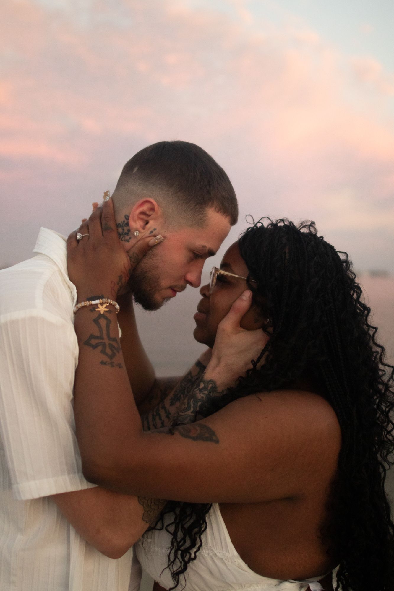 Tattooed couple in white outfits sharing a romantic moment under a pastel pink sunset sky.