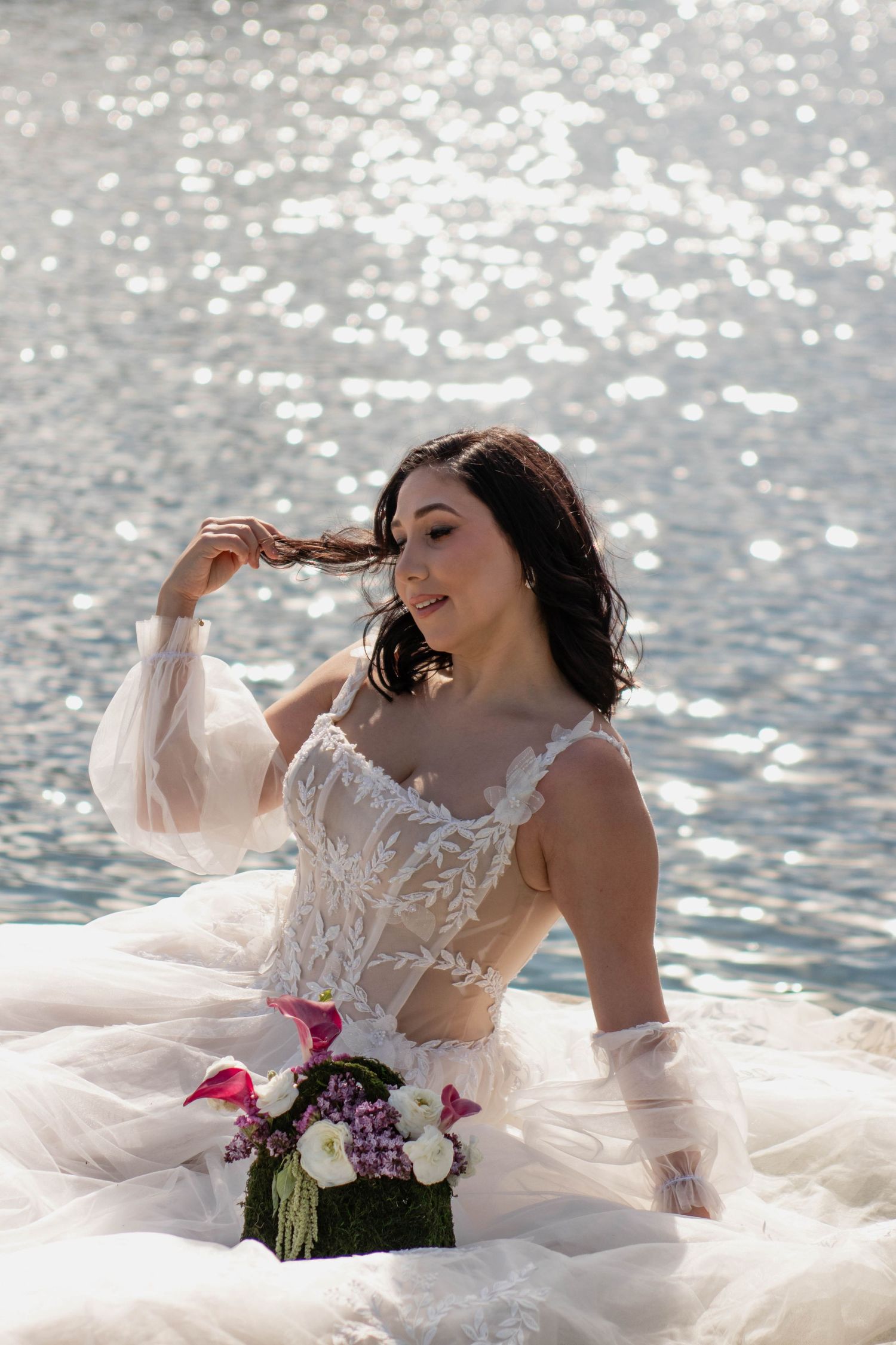 Brunette bride in lace wedding dress sitting by sparkling water, holding bouquet of pink flowers.