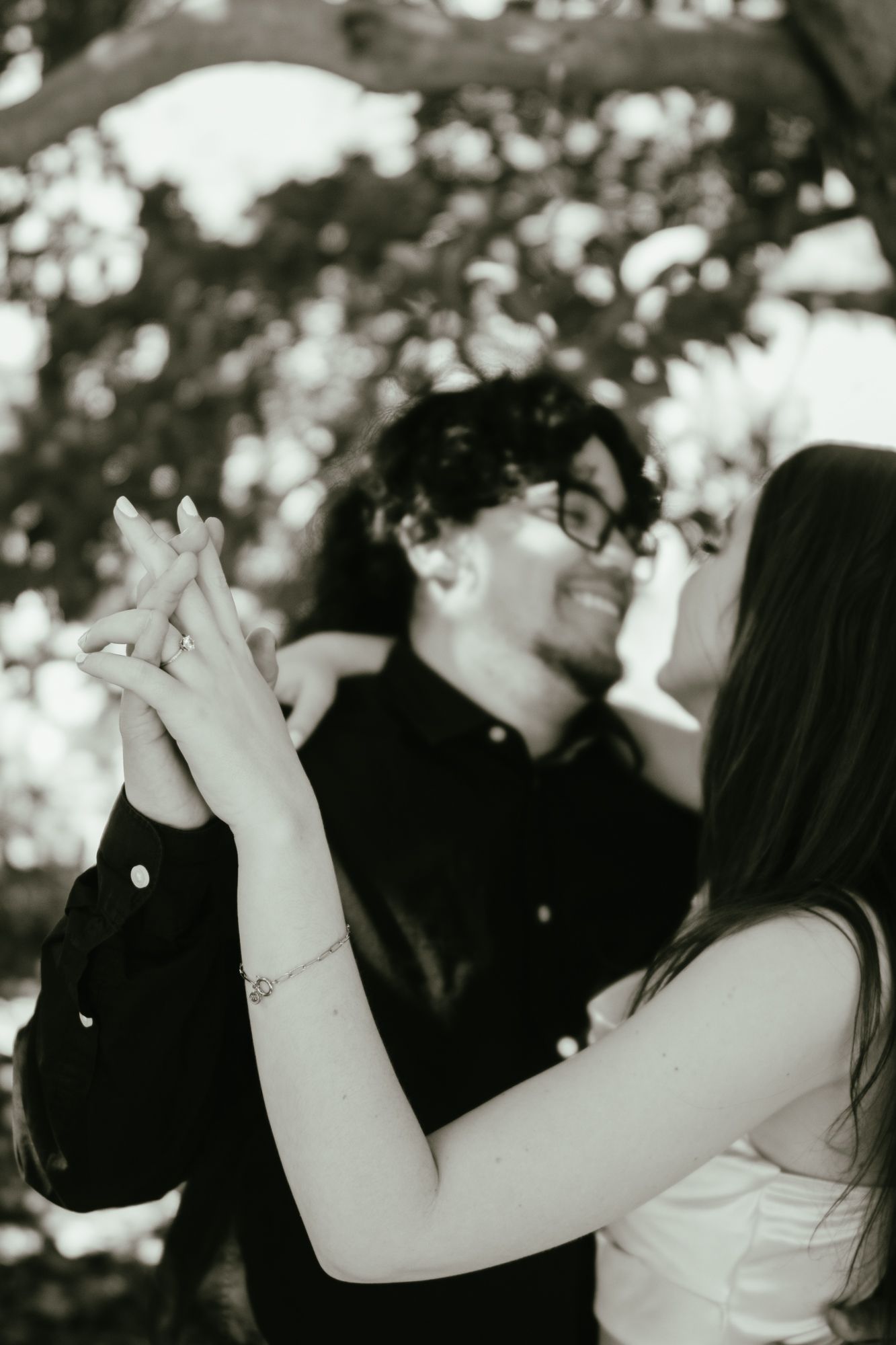 Black and white photo of engaged couple holding hands together outdoors under trees.