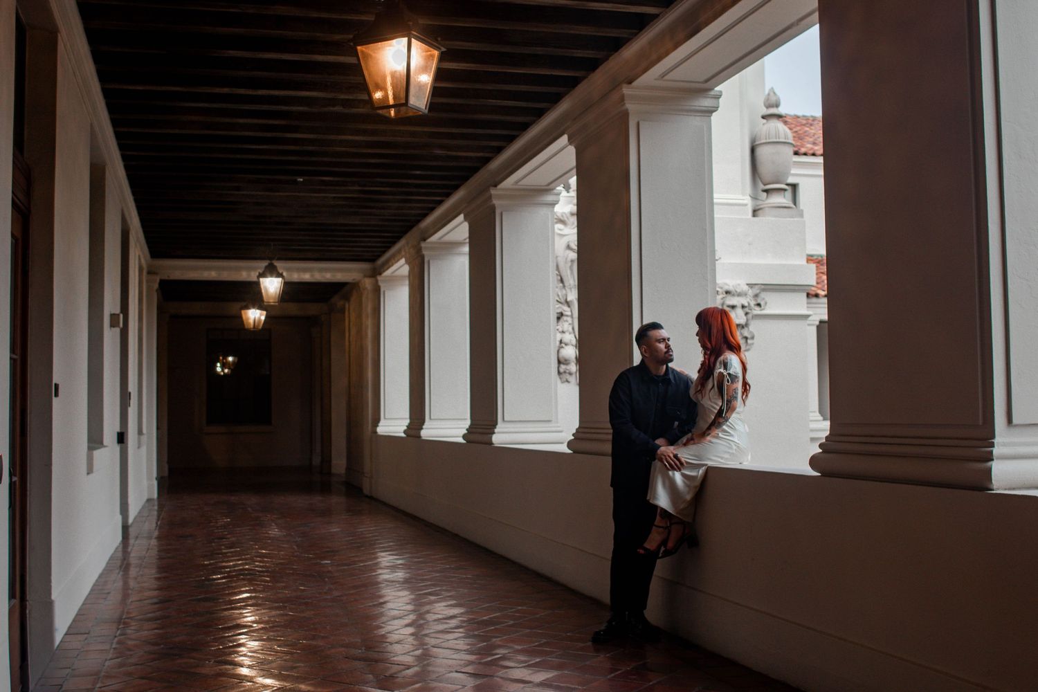 Couple sharing an intimate moment in a sunlit historic corridor with arched columns.