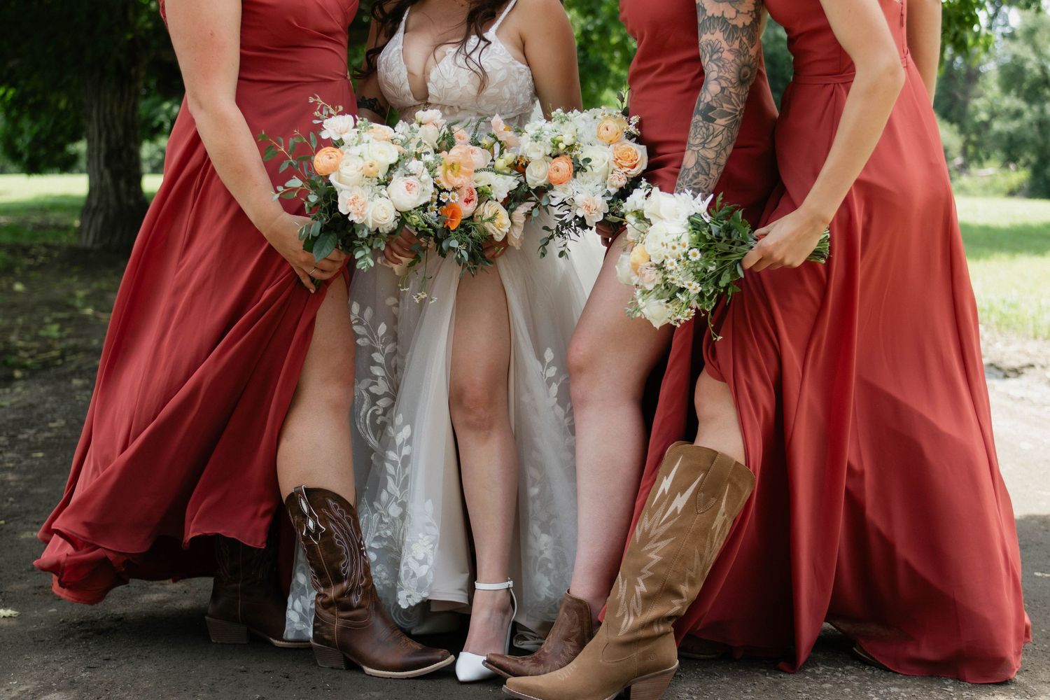 Bride and bridesmaids in red dresses wearing cowboy boots holding floral bouquets outdoors.