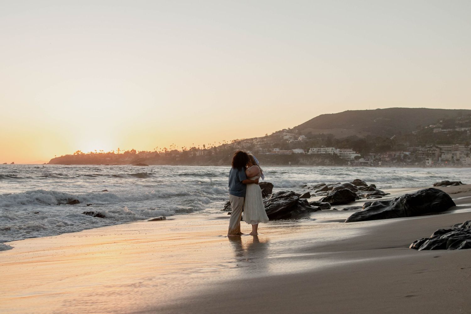 Couple embracing on a sandy beach at golden sunset with rocky shoreline and hills in the background.