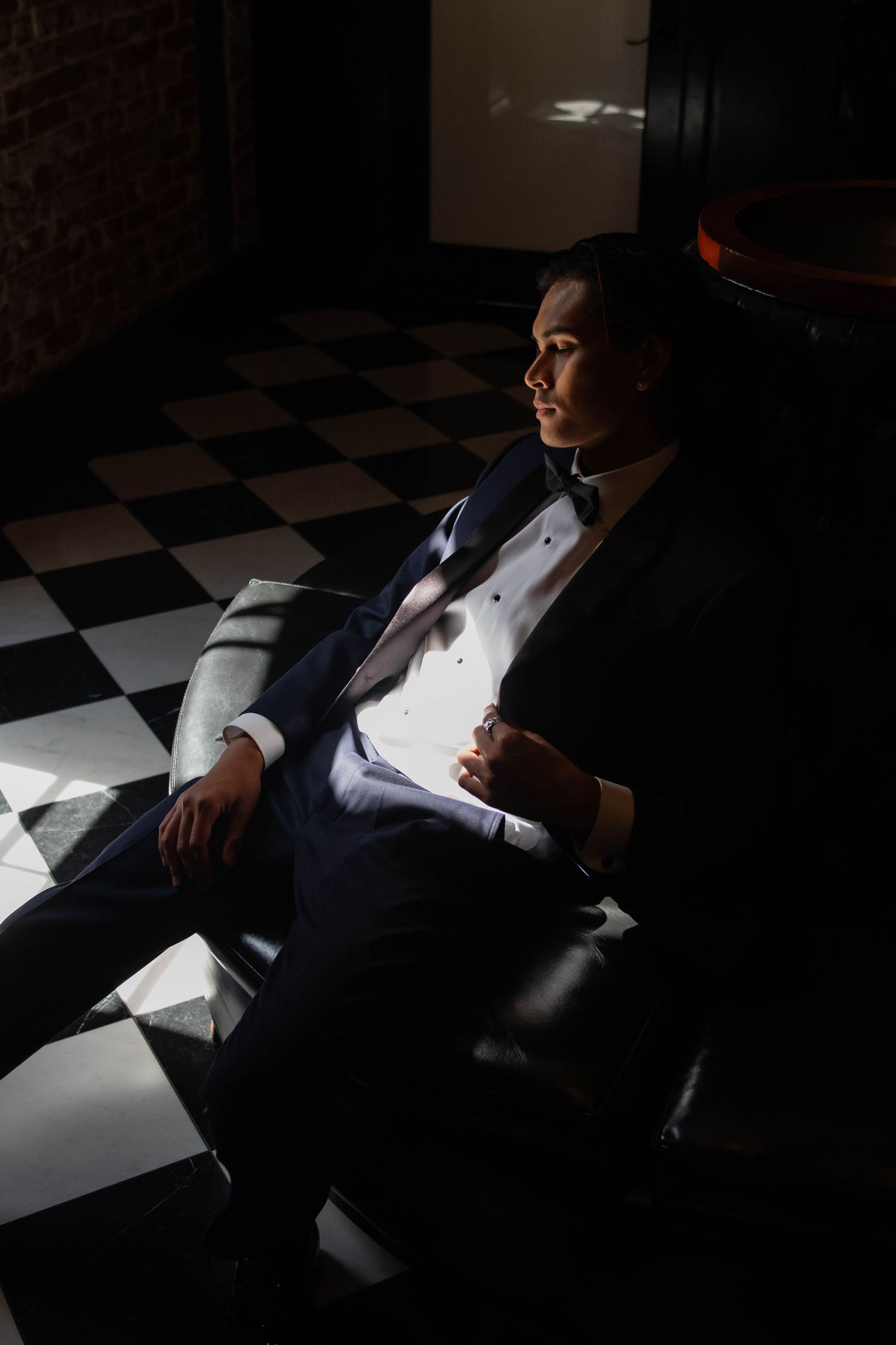 A groom in a tuxedo sits in a leather chair on a checkered floor, bathed in dramatic light.