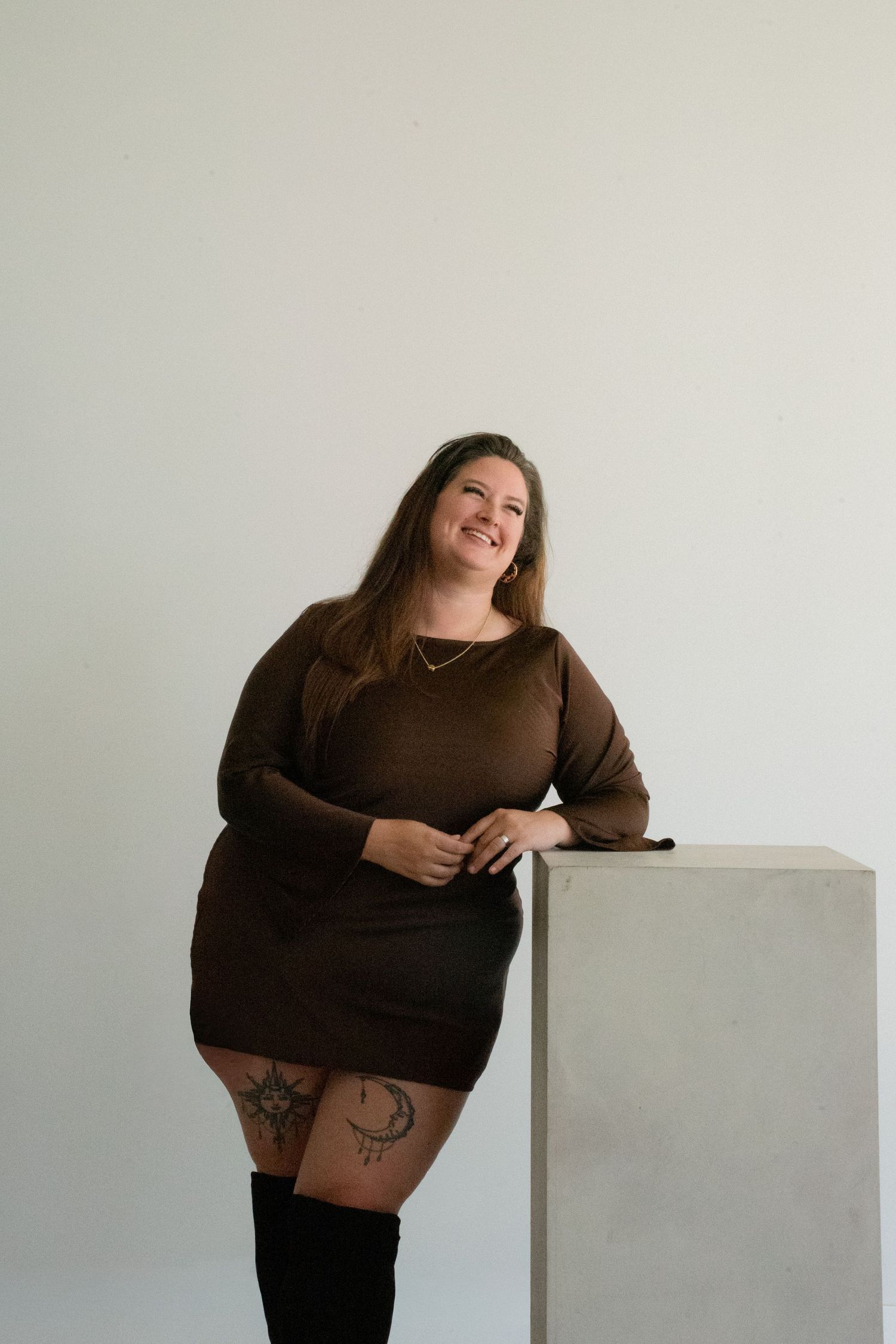 Confident plus size woman in brown sweater dress and thigh-high boots leans against white pedestal.