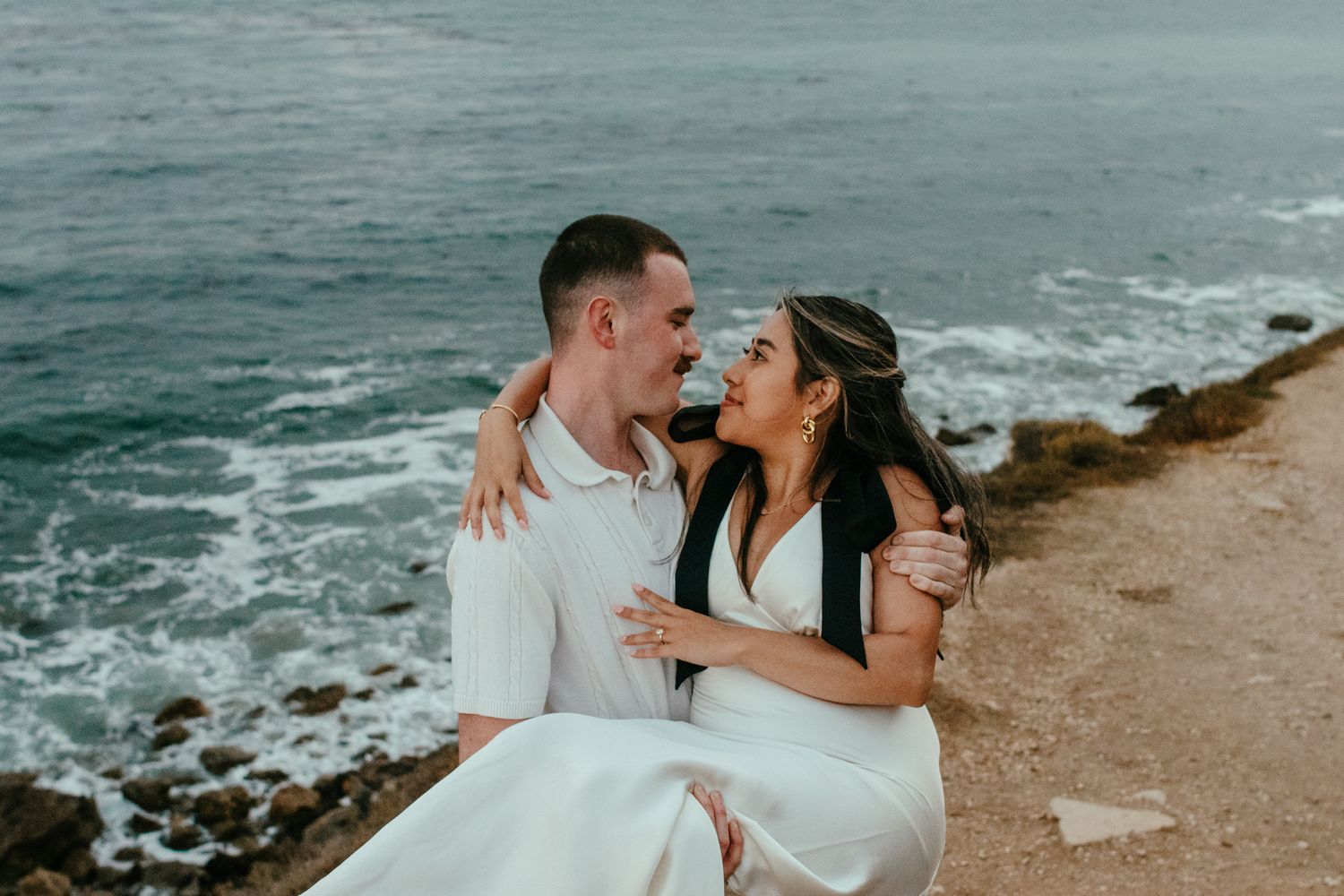 Man carries woman in white dress as they gaze lovingly at each other on scenic ocean cliff.