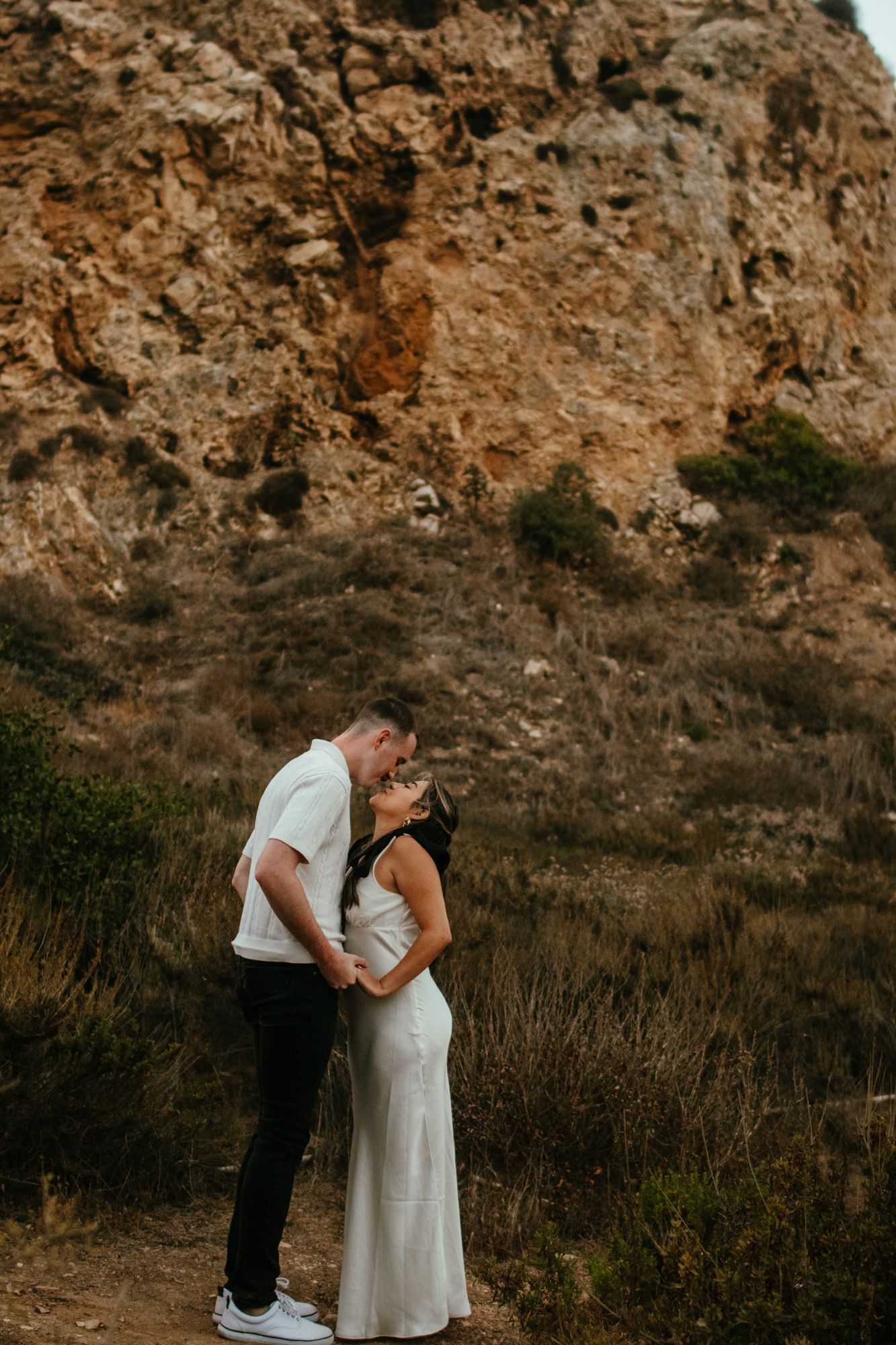 Couple sharing a tender kiss in front of a rugged rocky hillside during golden hour outdoors.