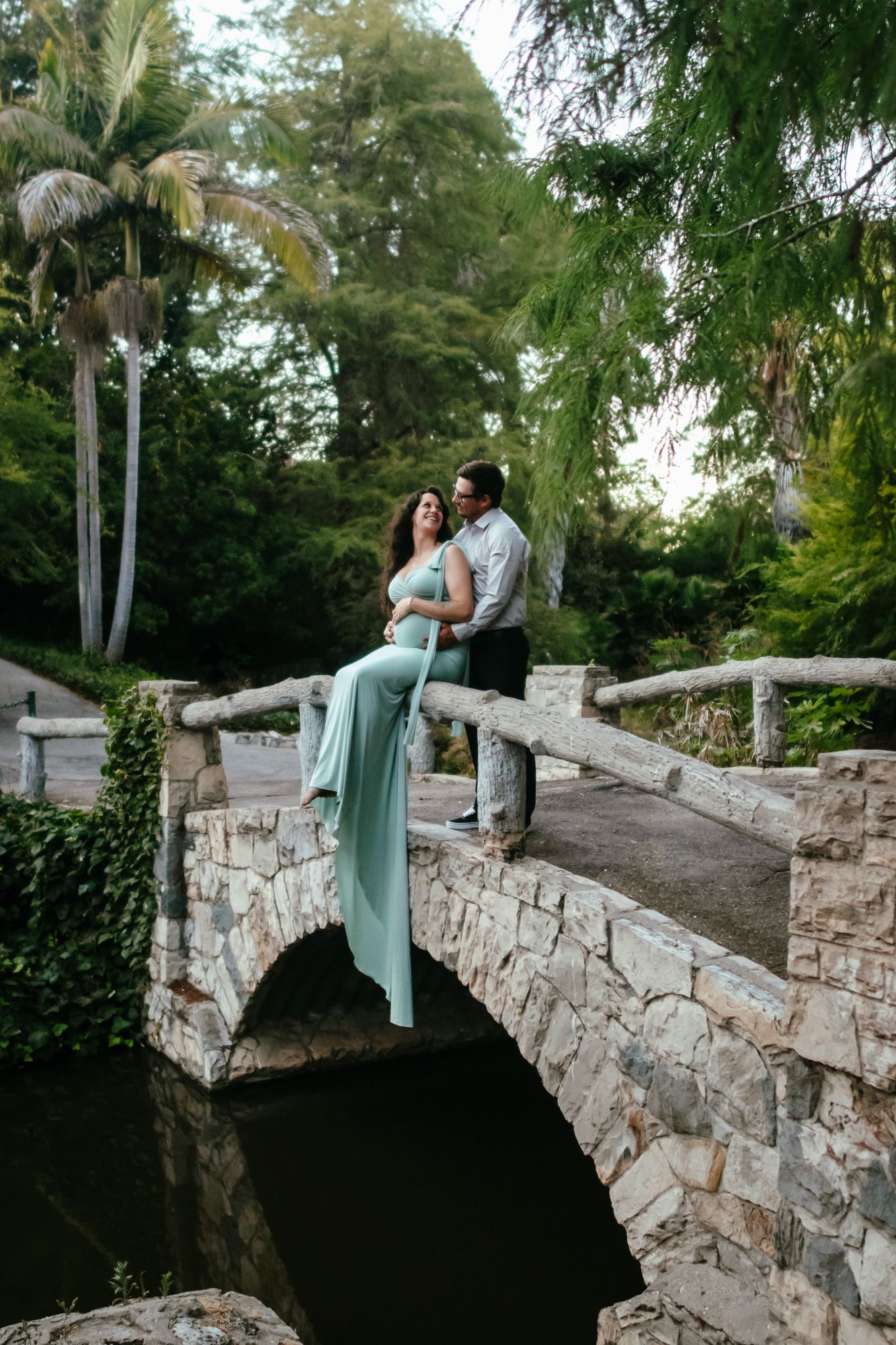 Expecting couple posing on a stone arch bridge surrounded by lush tropical garden greenery.