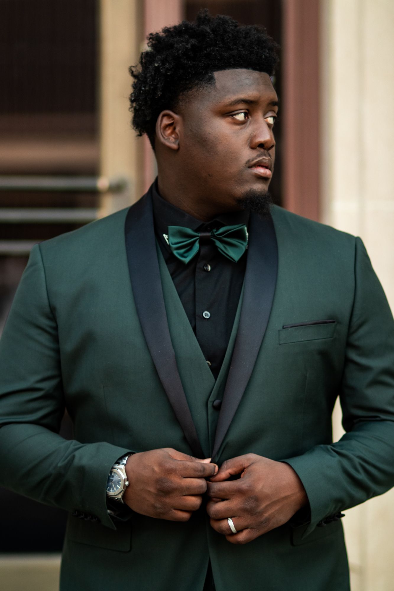 Groom in green tuxedo with black bow tie adjusting jacket button outdoors.