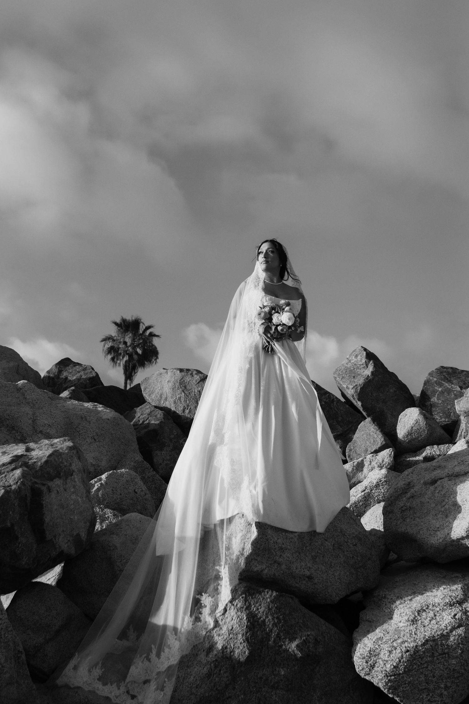 Bride in white gown and veil stands on rocky shore holding bouquet with palm tree and cloudy sky backdrop.