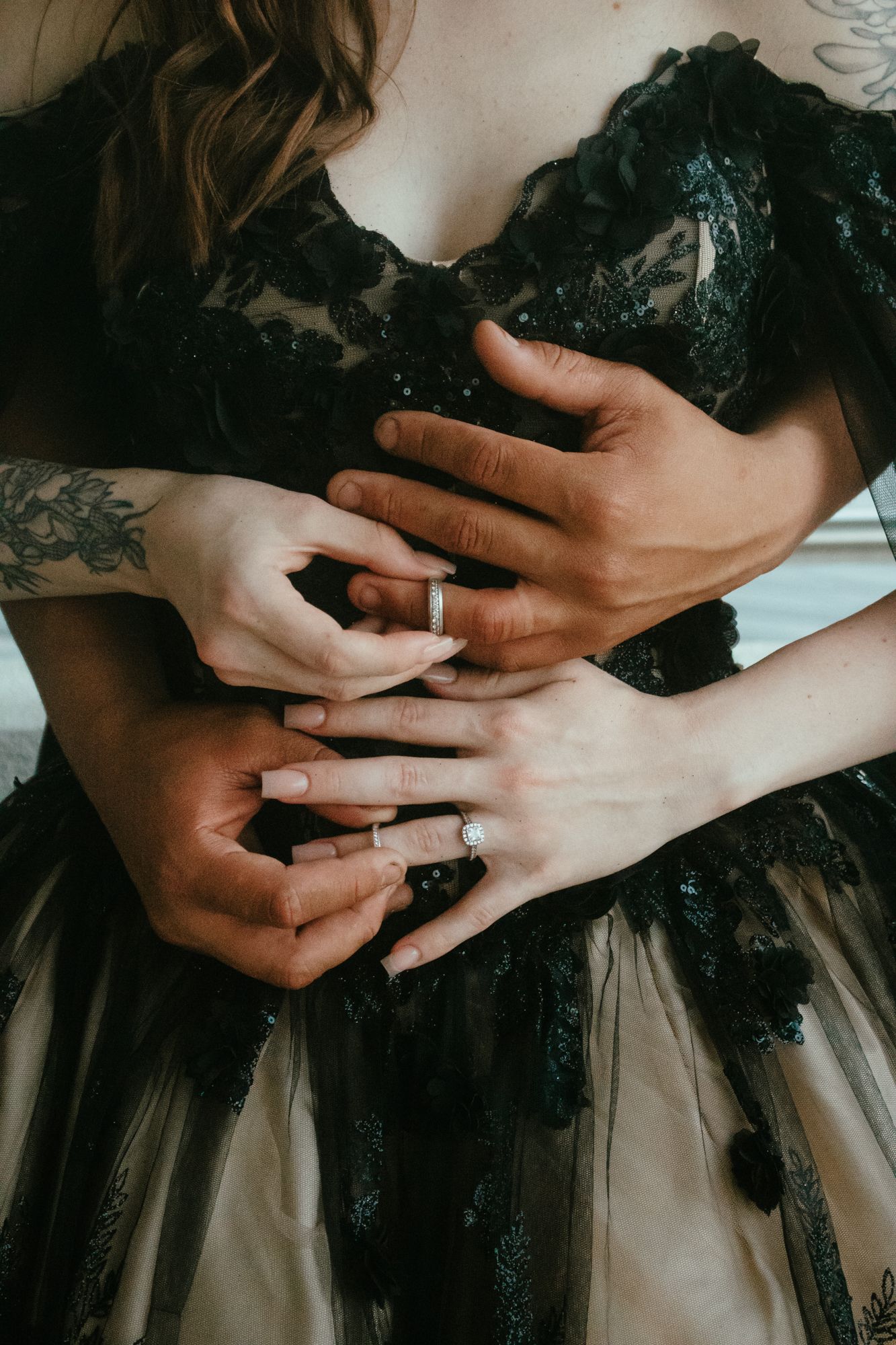 Couple embracing in black lace wedding gown, showing matching wedding bands and tattooed hands.