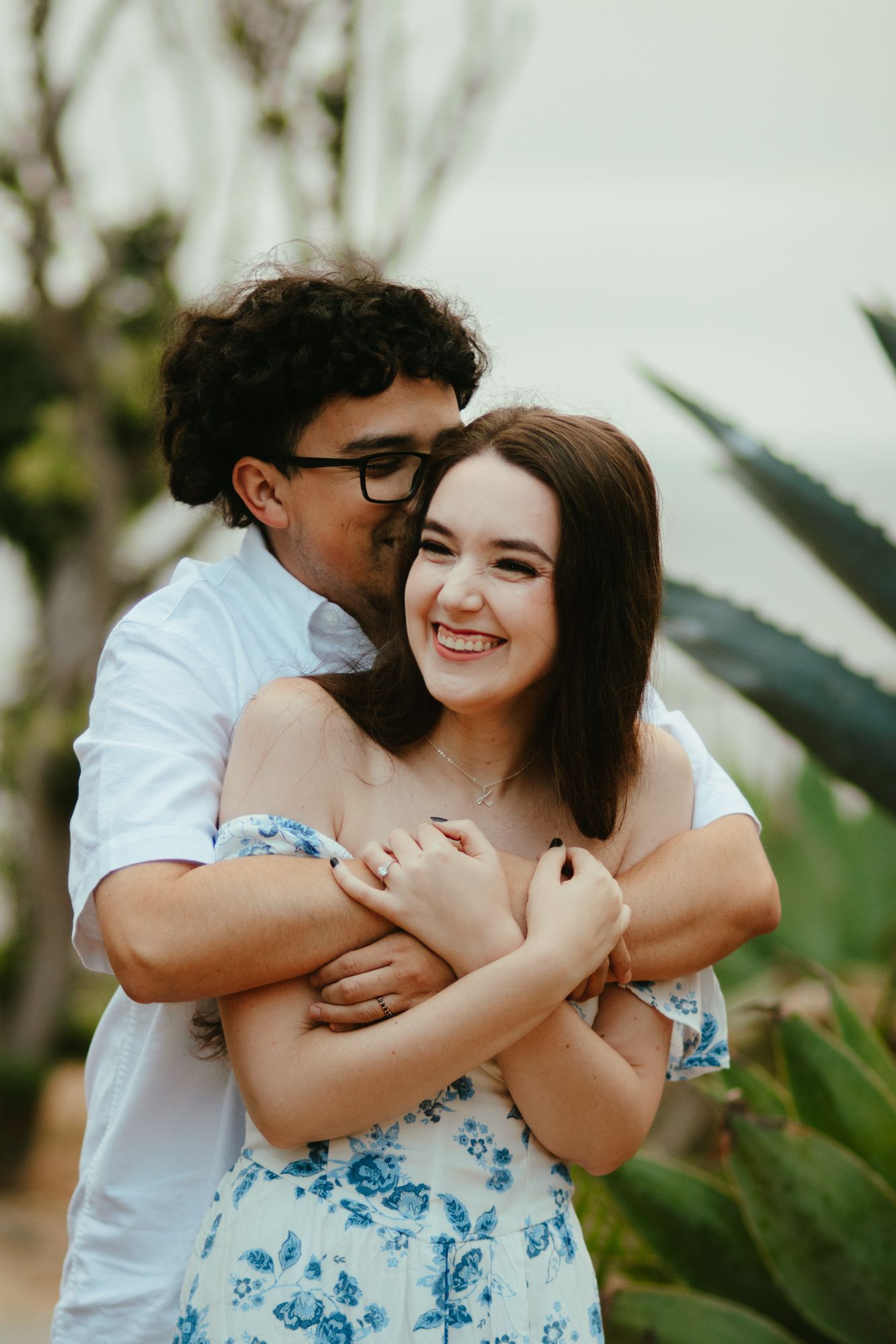 Man hugging laughing woman from behind near agave plants in casual outdoor engagement photo.