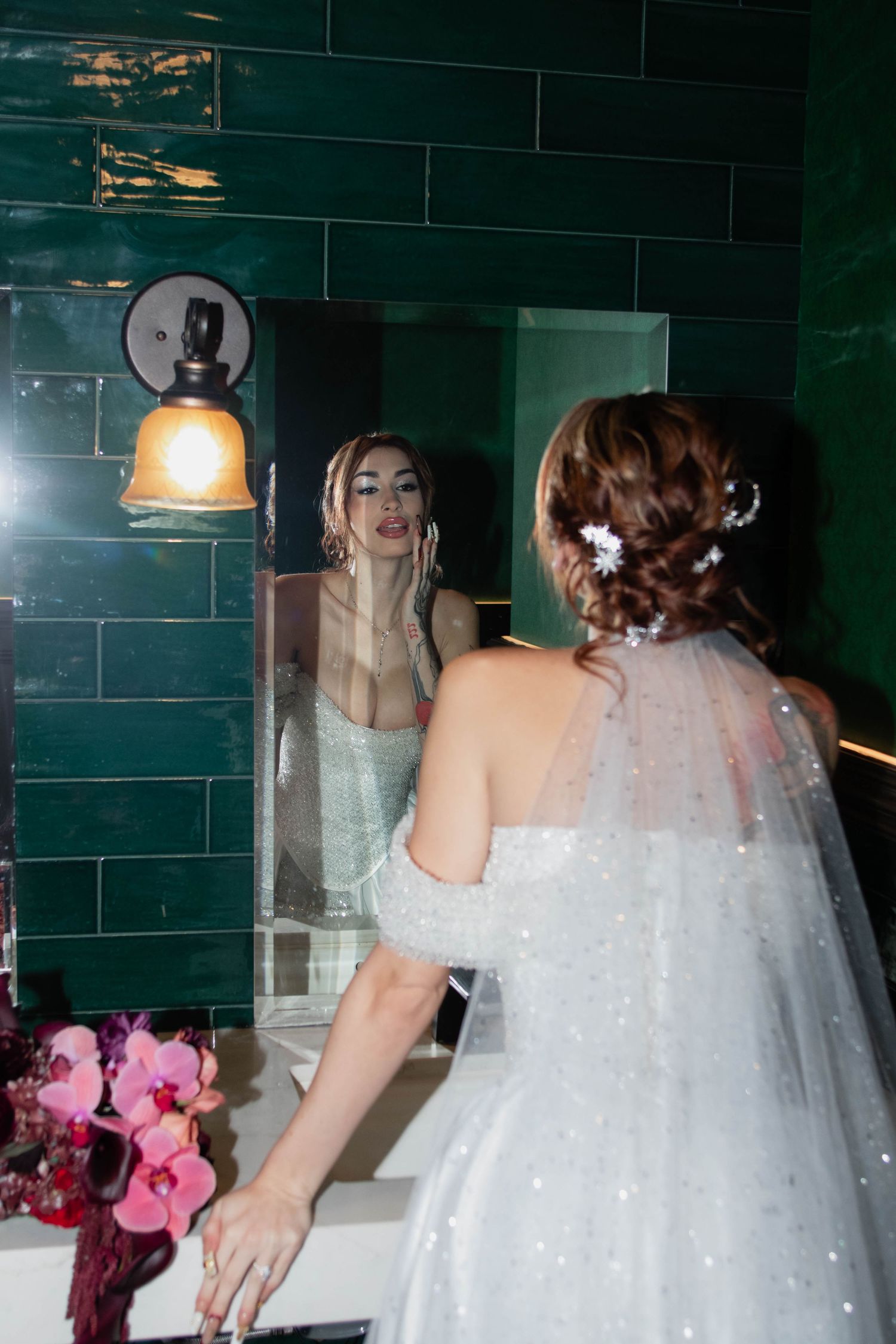 Bride in white wedding gown with floral hair accessories checks her reflection in mirror with green tile wall.