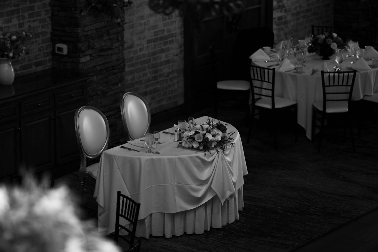 Black and white photo of elegant wedding reception sweetheart table with floral centerpiece in brick venue.