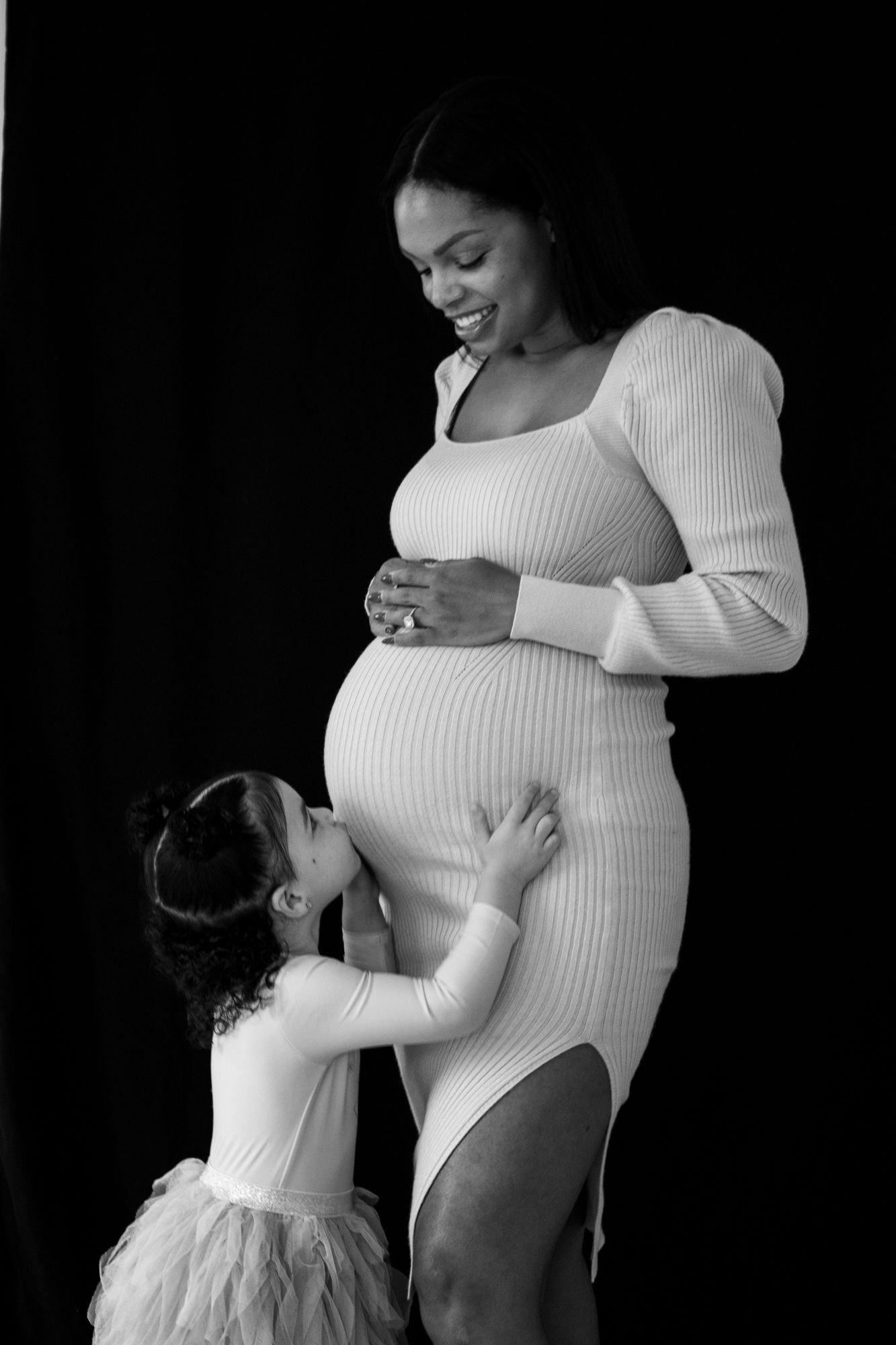 Black and white photo of smiling pregnant woman in fitted dress as young daughter kisses her baby bump.