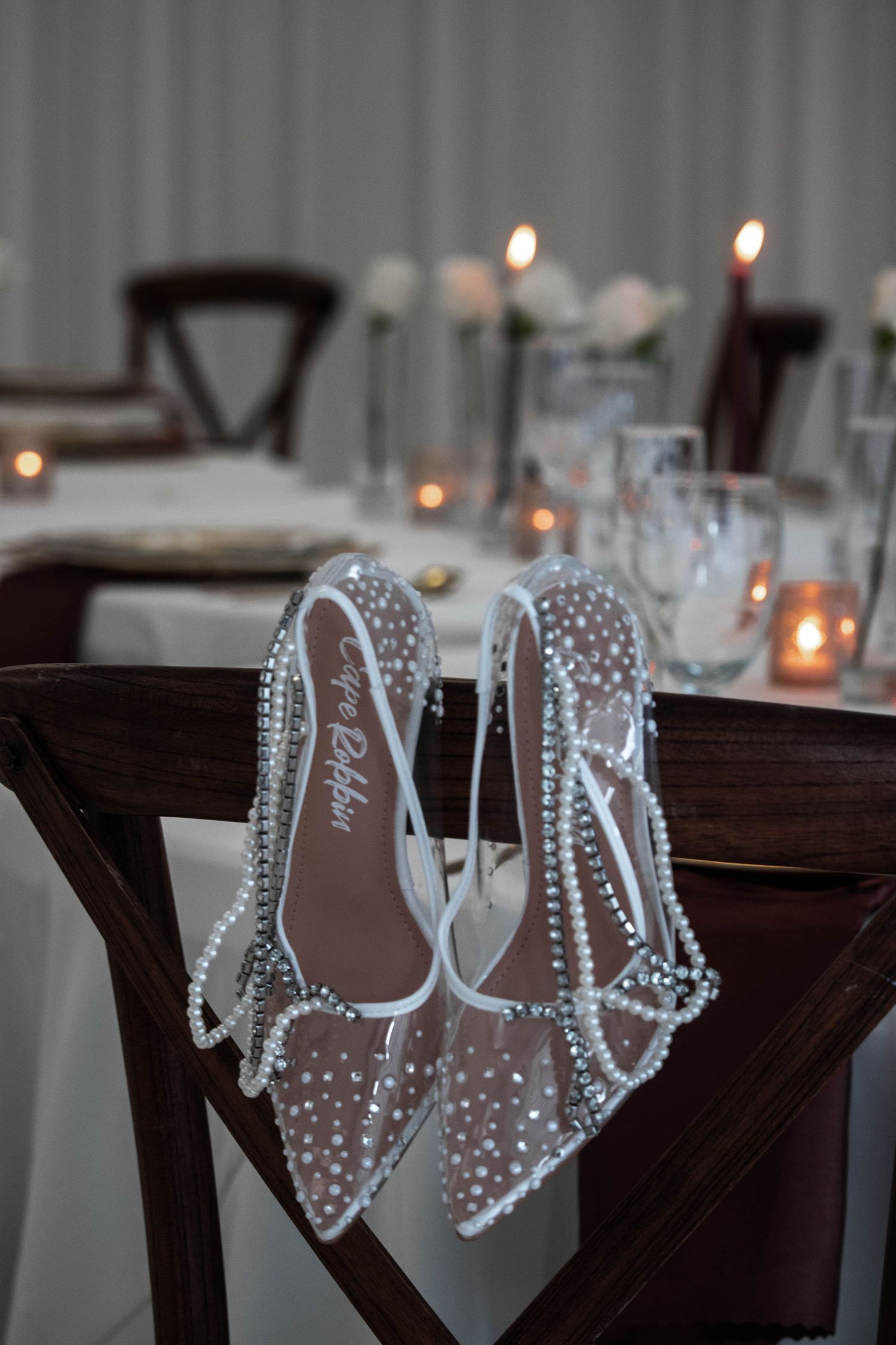 Crystal-embellished bridal heels by Amina Muaddi displayed on a dark wooden chair with candlelit reception tables behind.