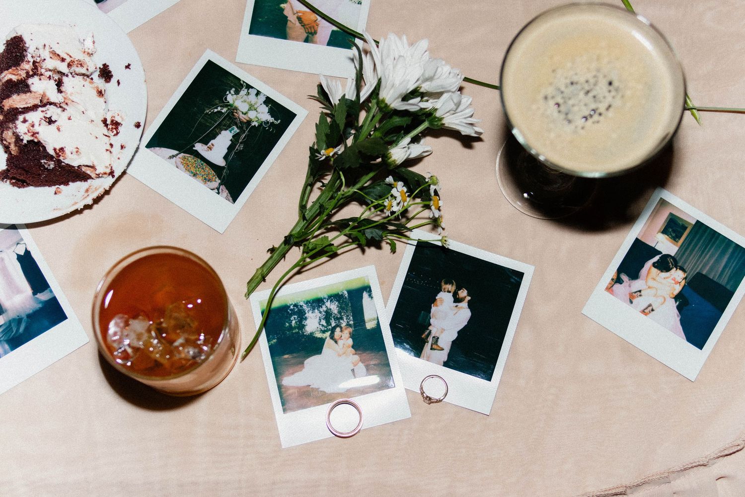 Flat lay of Polaroid wedding photos, rings, flowers, coffee, and drinks on a wooden table.