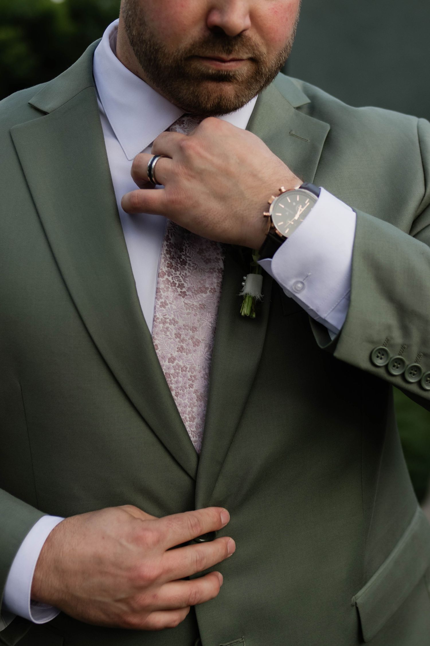 Groom in sage green suit adjusting floral tie with boutonniere and watch detail close-up.