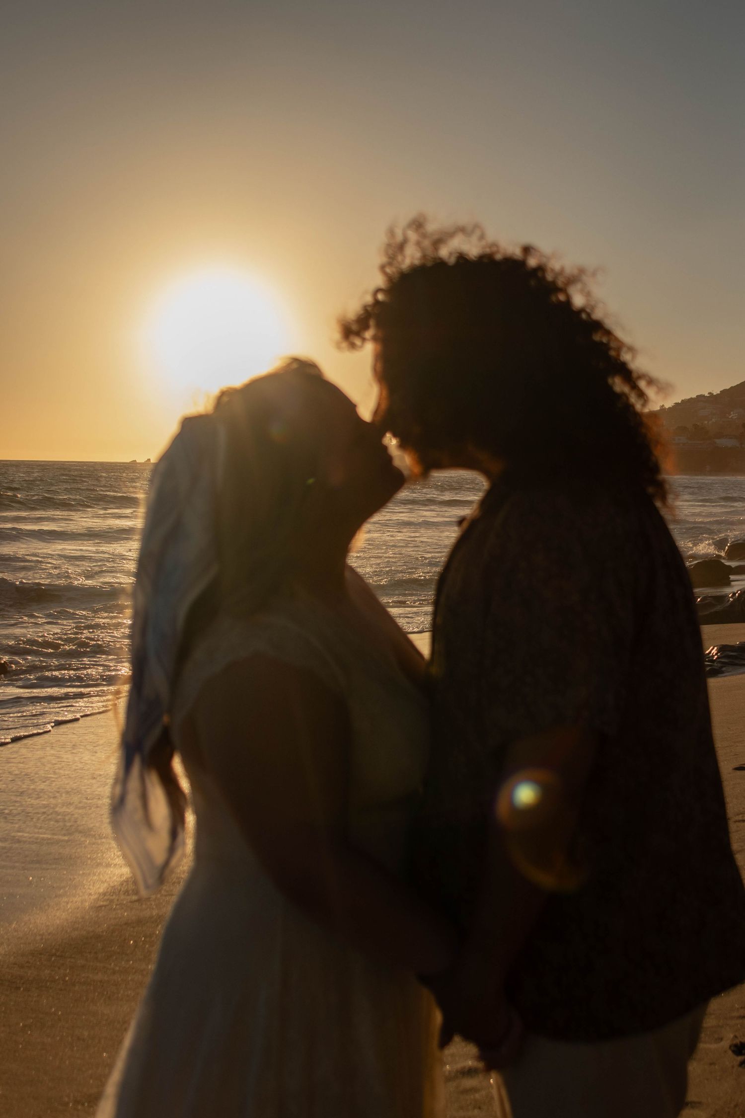 Couple sharing a romantic kiss on the beach at golden sunset, bride in white veil, ocean waves in background.