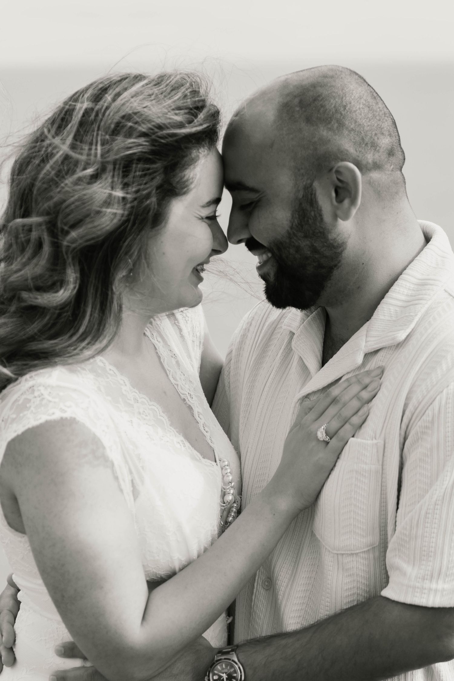 Black and white portrait of laughing couple touching foreheads with ocean in the background.
